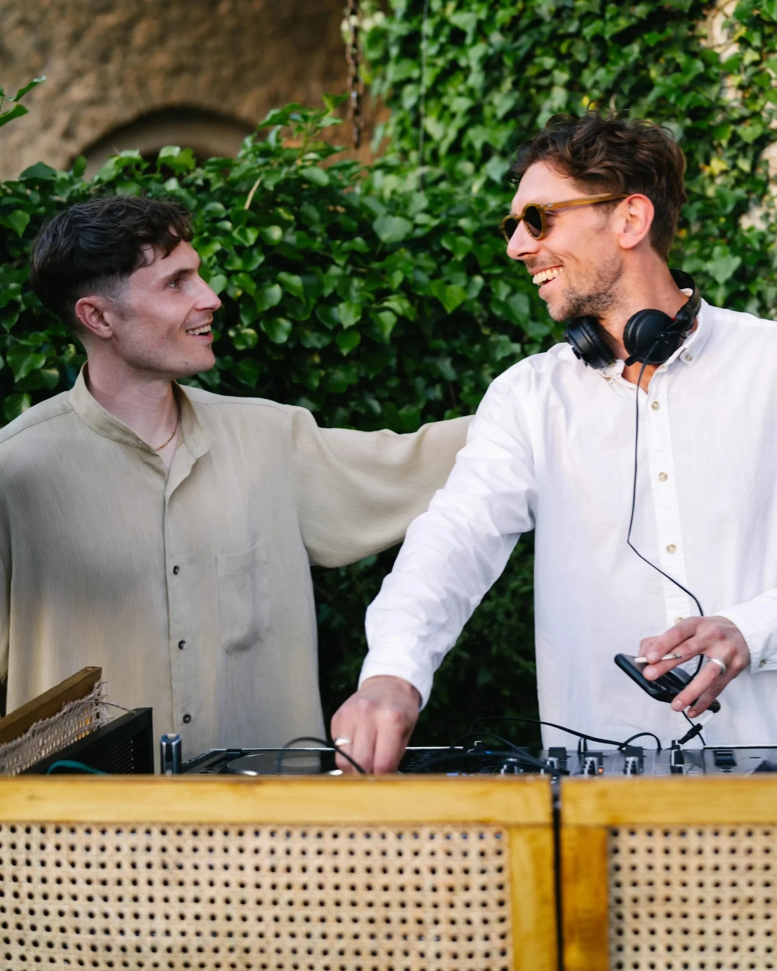 Two men smiling and greeting each other at an outdoor event, with DJ equipment and greenery in the background.