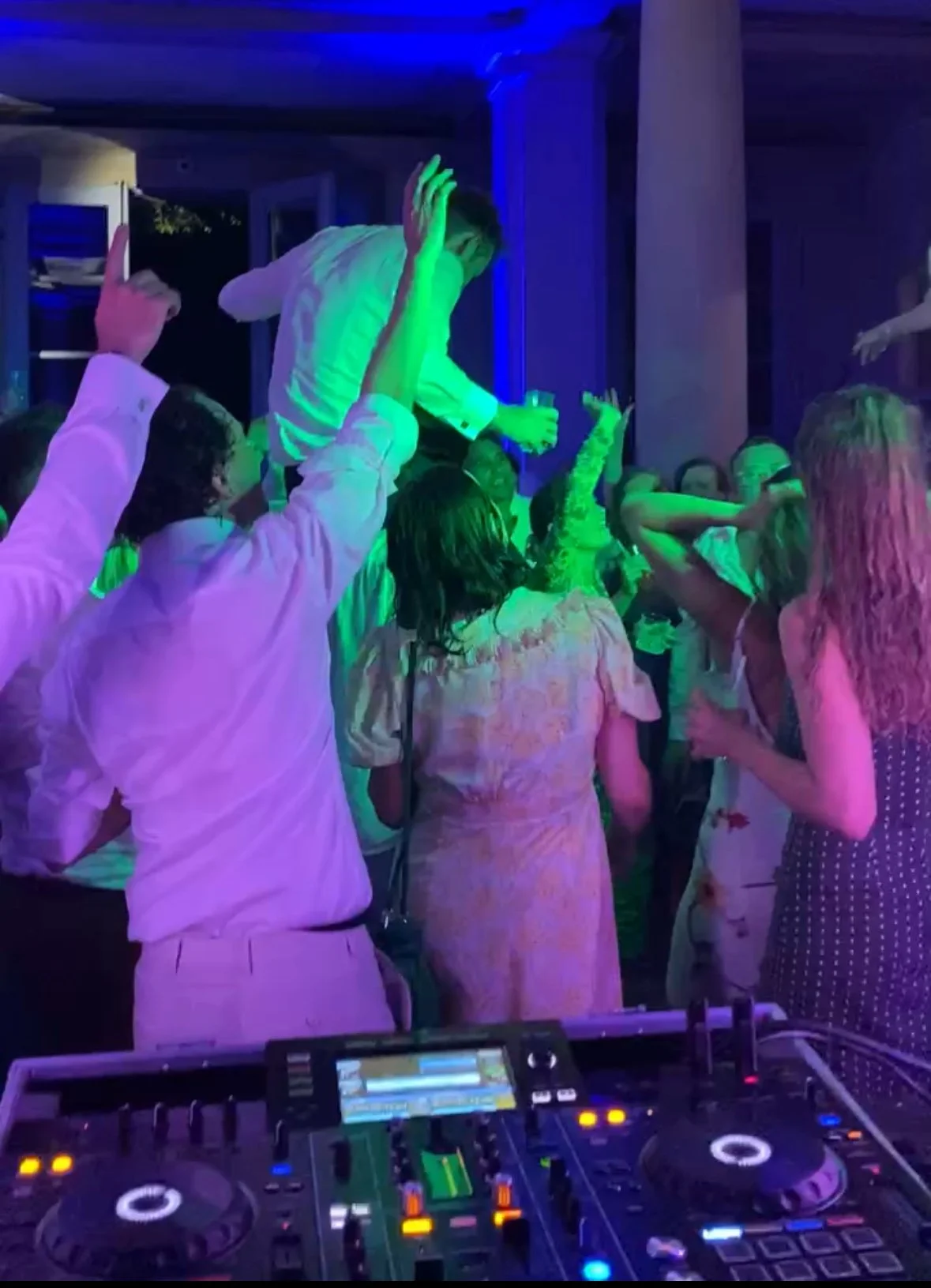 People dancing at a party with colorful lighting, DJ equipment in the foreground, some individuals raising hands, and a child being lifted over others