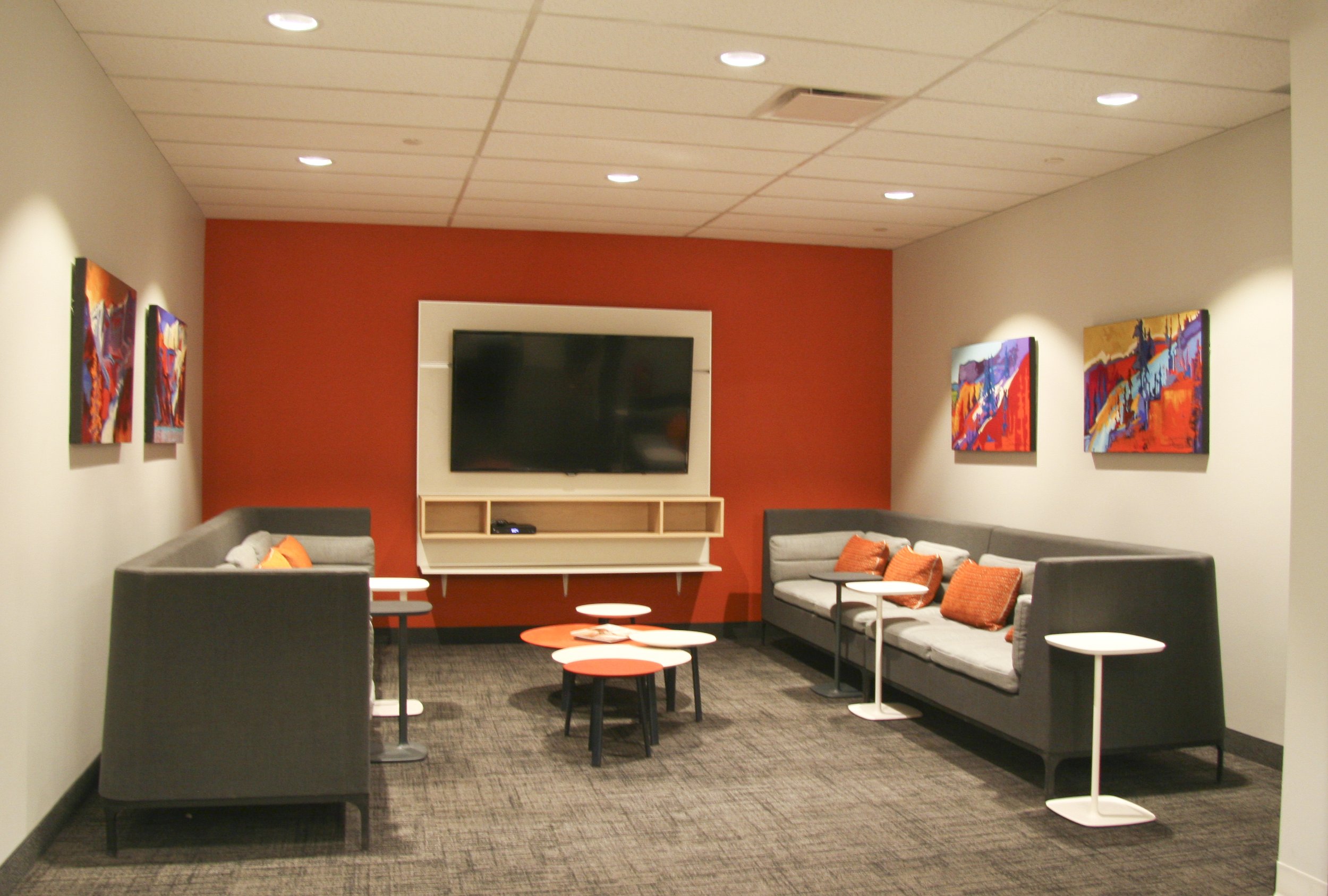 Modern lounge area with grey couches, colorful abstract paintings, a TV on a red accent wall, and small white tables.