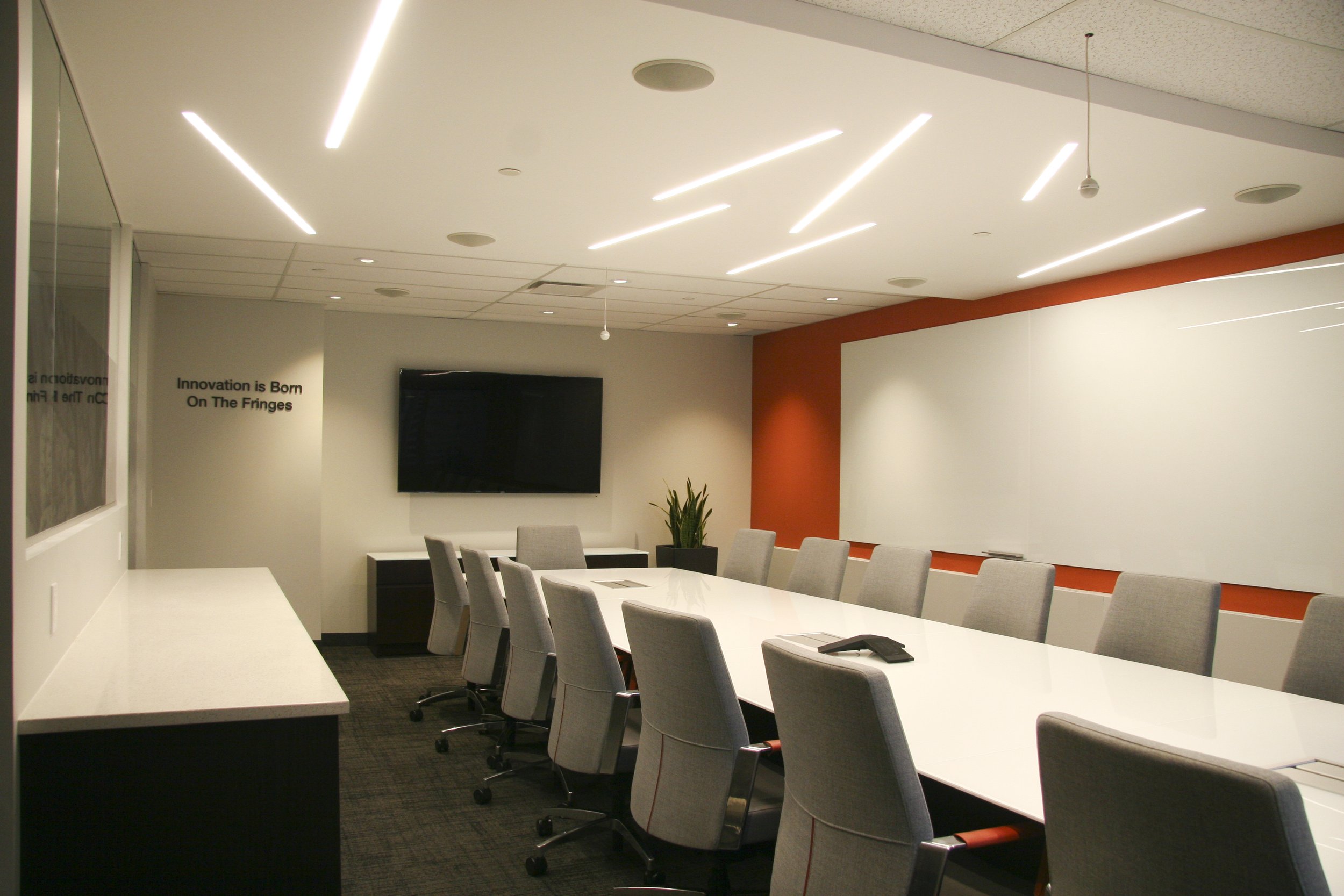 Modern conference room with a long white table, gray chairs, large screen, and minimalist ceiling lights. Whiteboard on orange accent wall.
