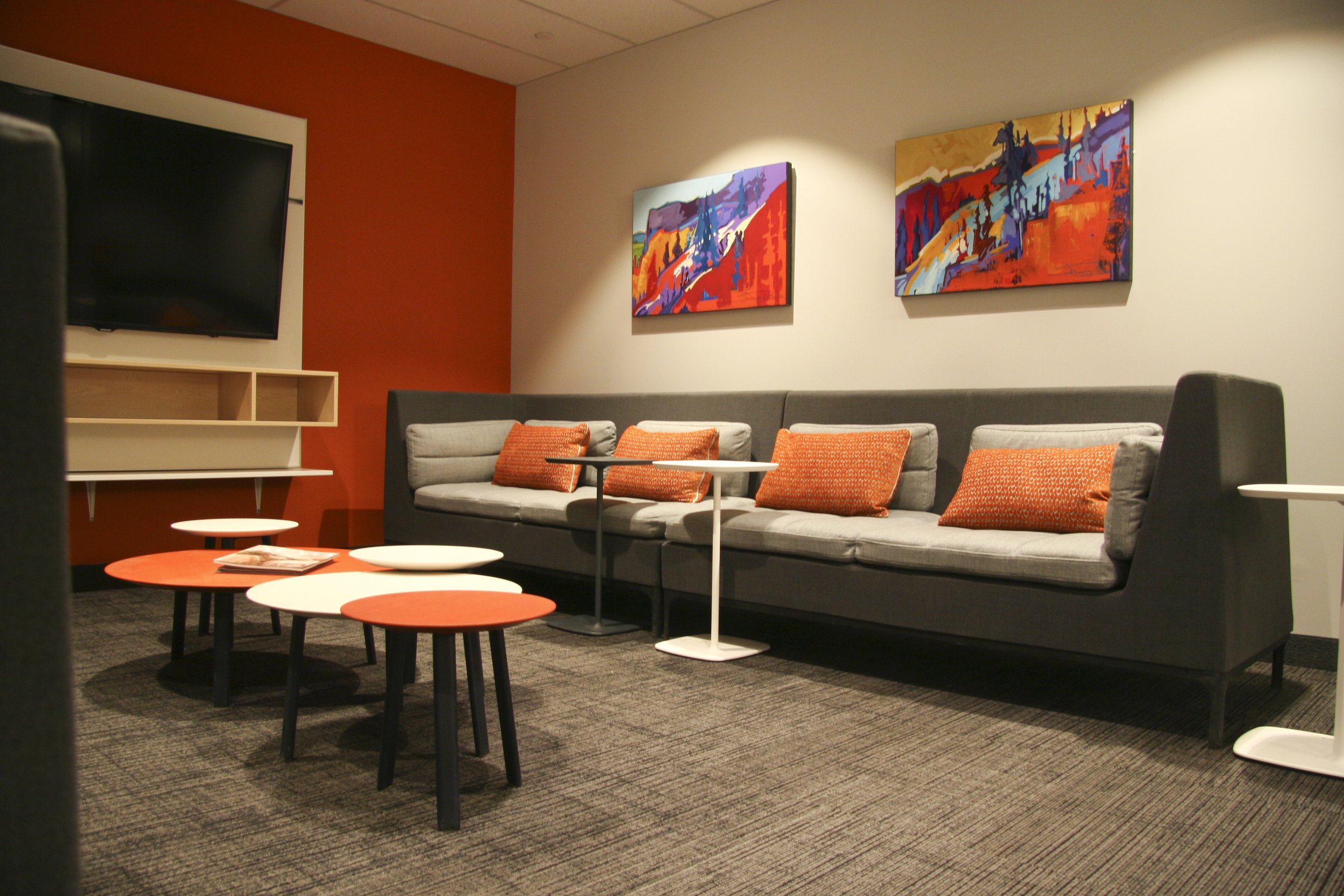 Modern lounge area with gray sofa, orange cushions, abstract wall art, round tables, TV, and orange accent wall.