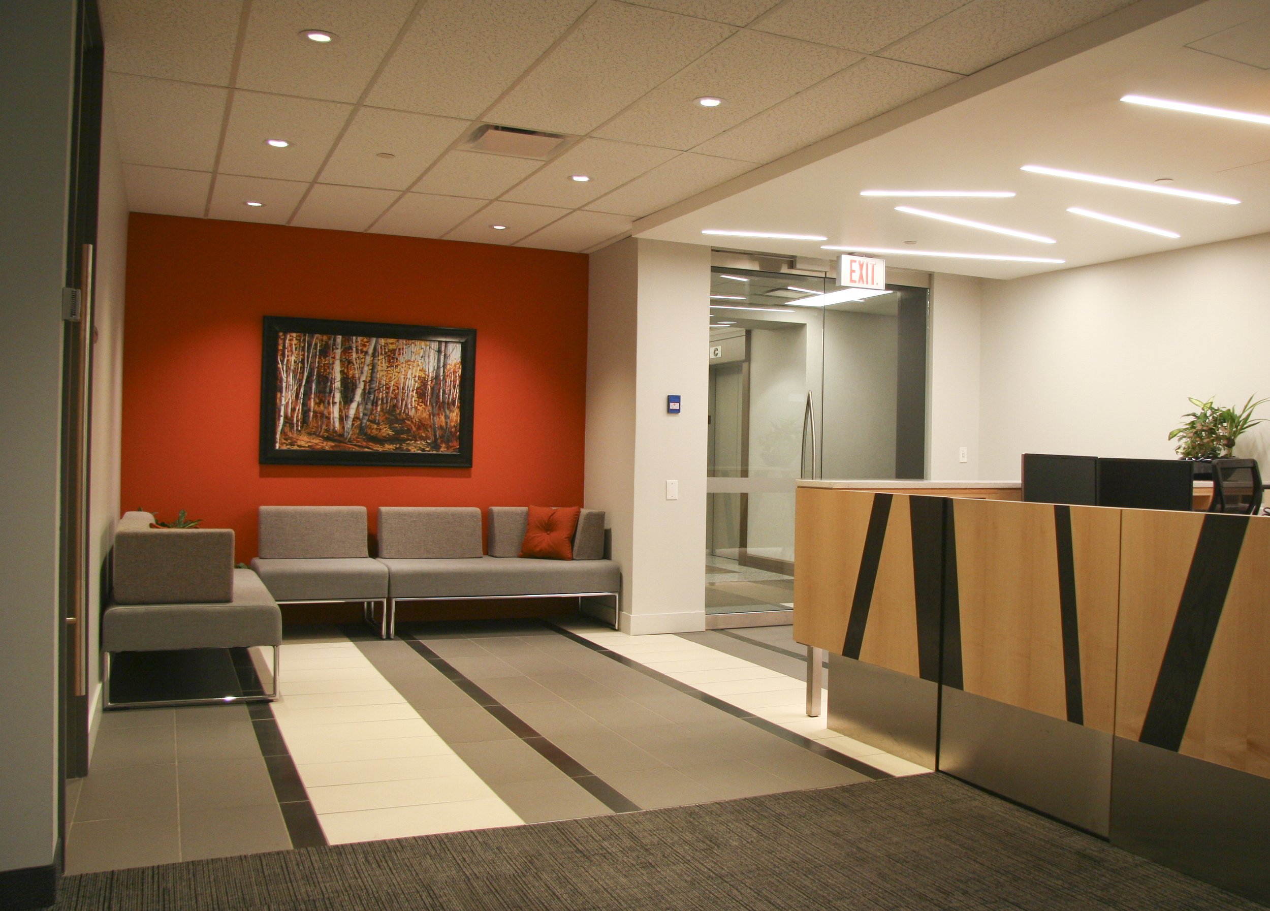 Modern office reception area with a red accent wall, gray seating, artwork, and a wooden front desk.