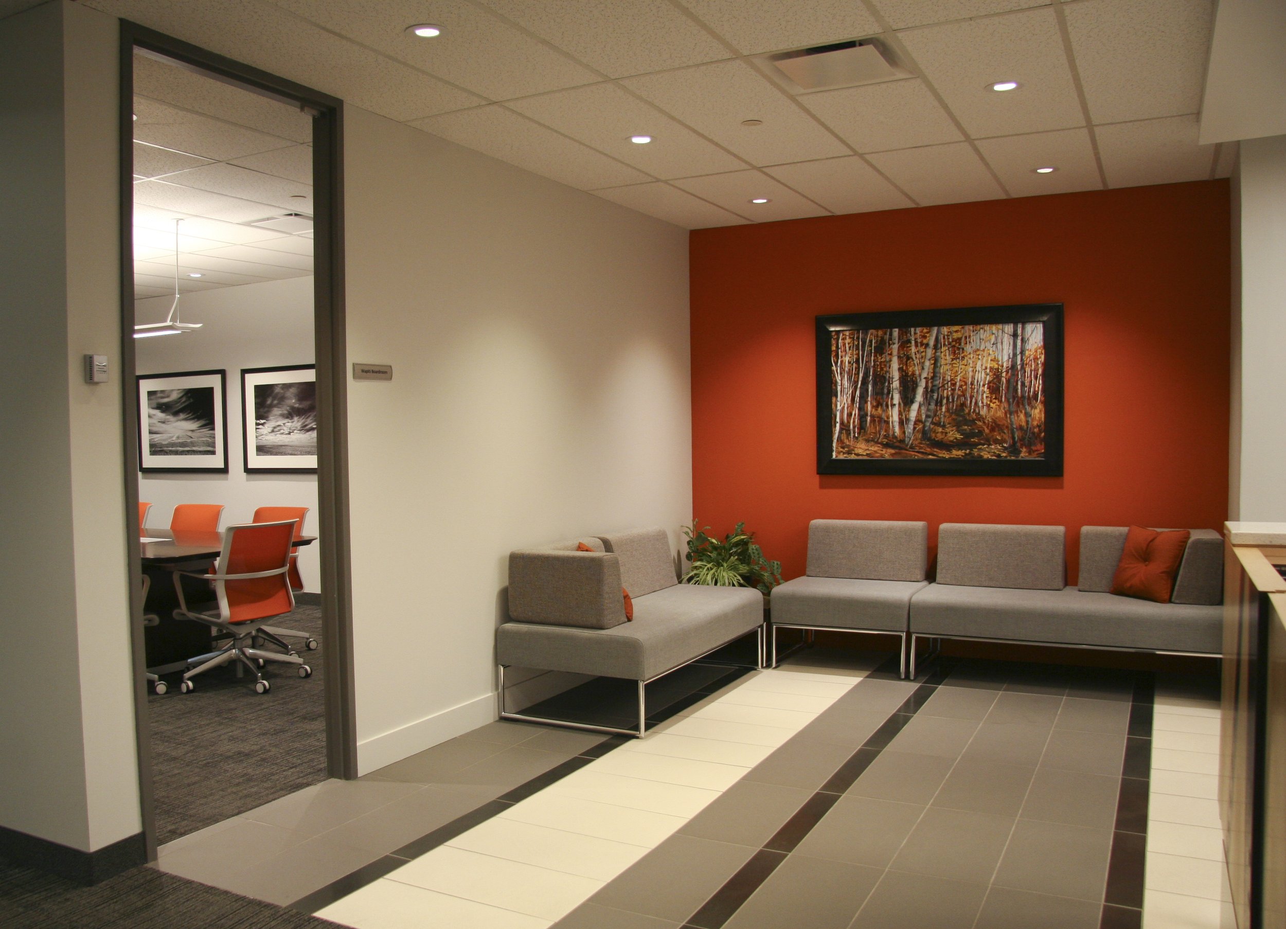 Office lobby with gray couches, orange accent wall, framed forest artwork, tiled floor, and adjacent conference room with orange chairs.
