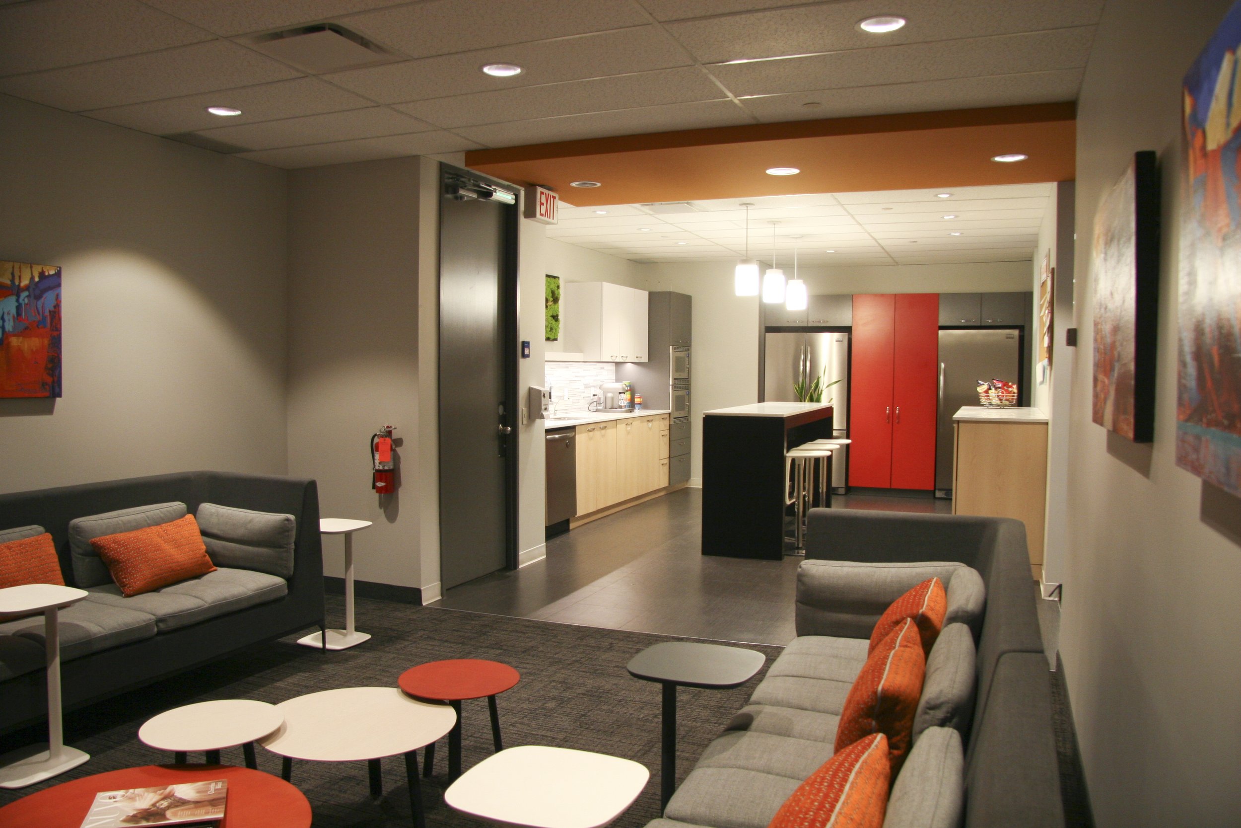 Modern office lounge area with gray couches and orange cushions, small round tables, kitchenette with cabinets, fridge, and bar seating.