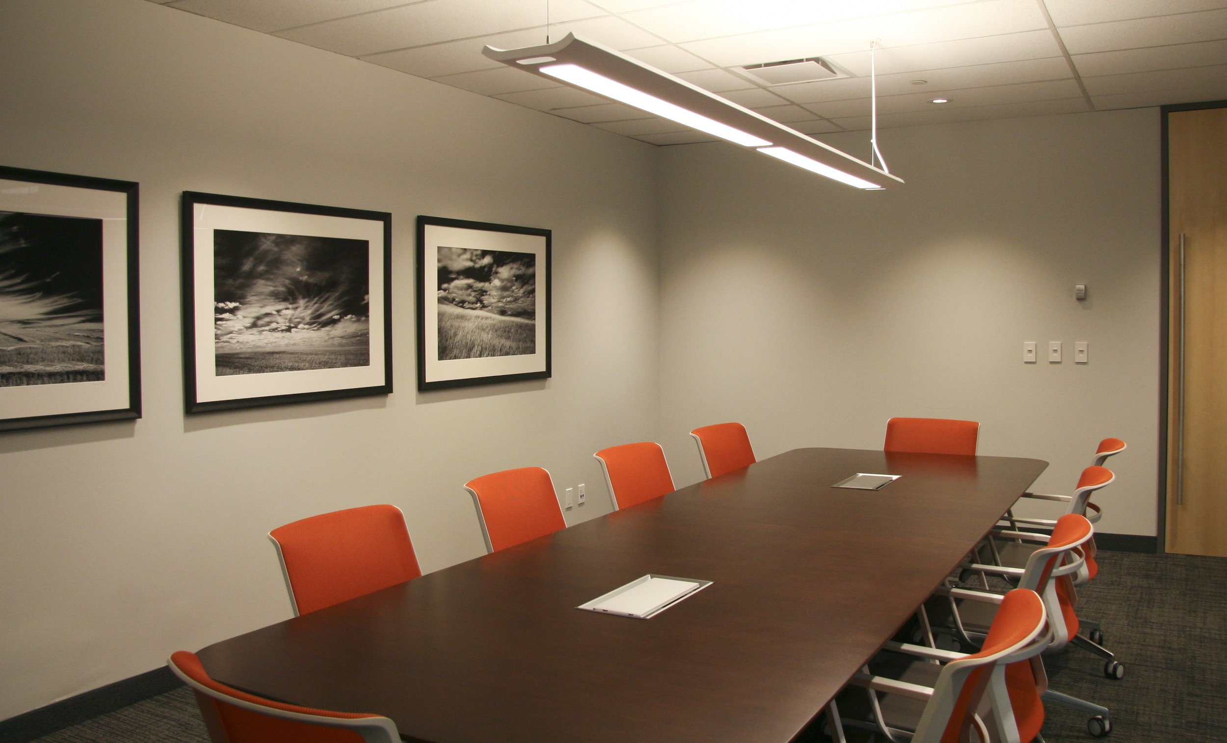 Conference room with long wooden table and orange chairs, three monochrome landscape photos on the wall, ceiling light above.