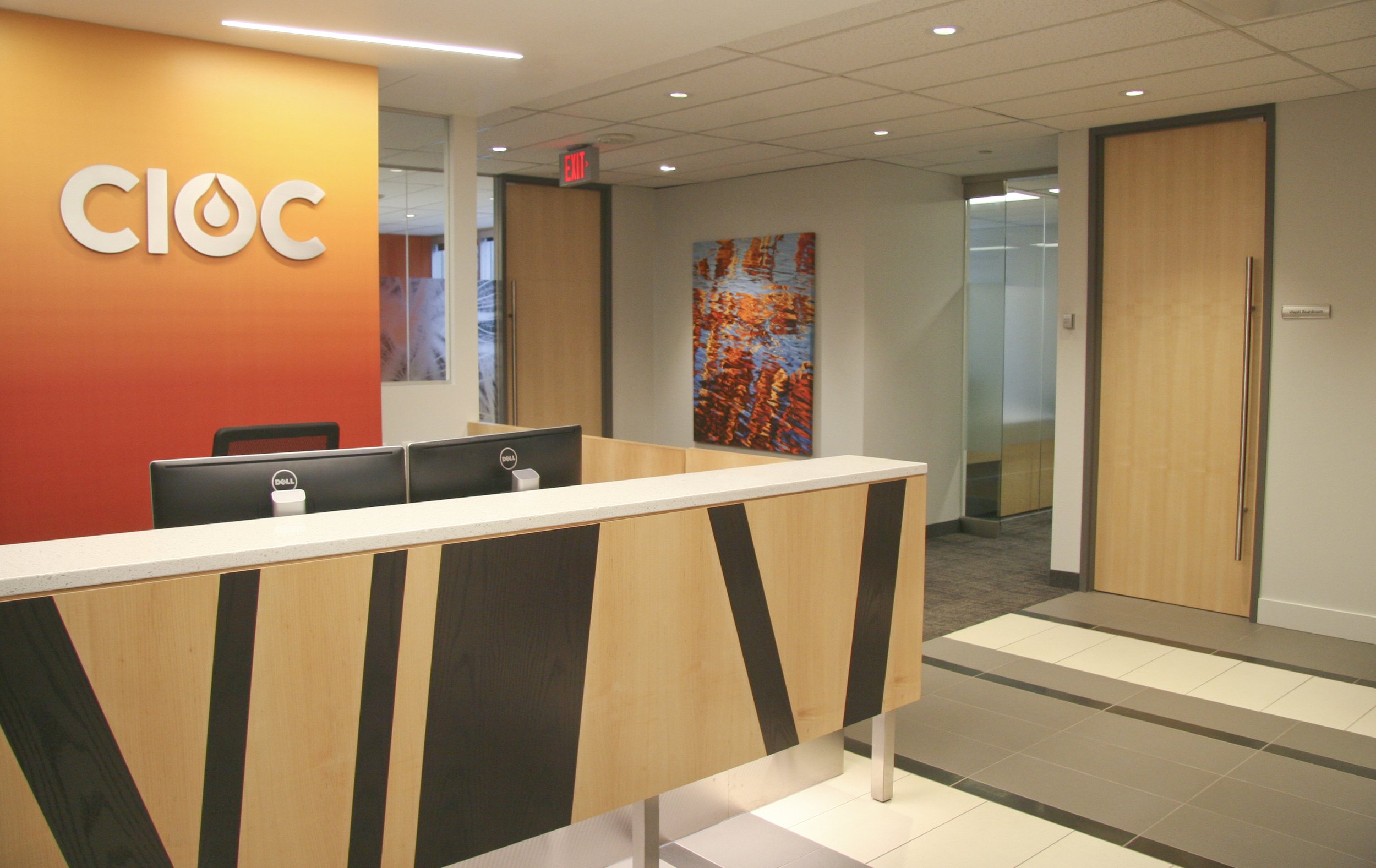 Modern office reception area with CIOC logo on orange wall, front desk with two Dell monitors, abstract art piece, and a wooden door.