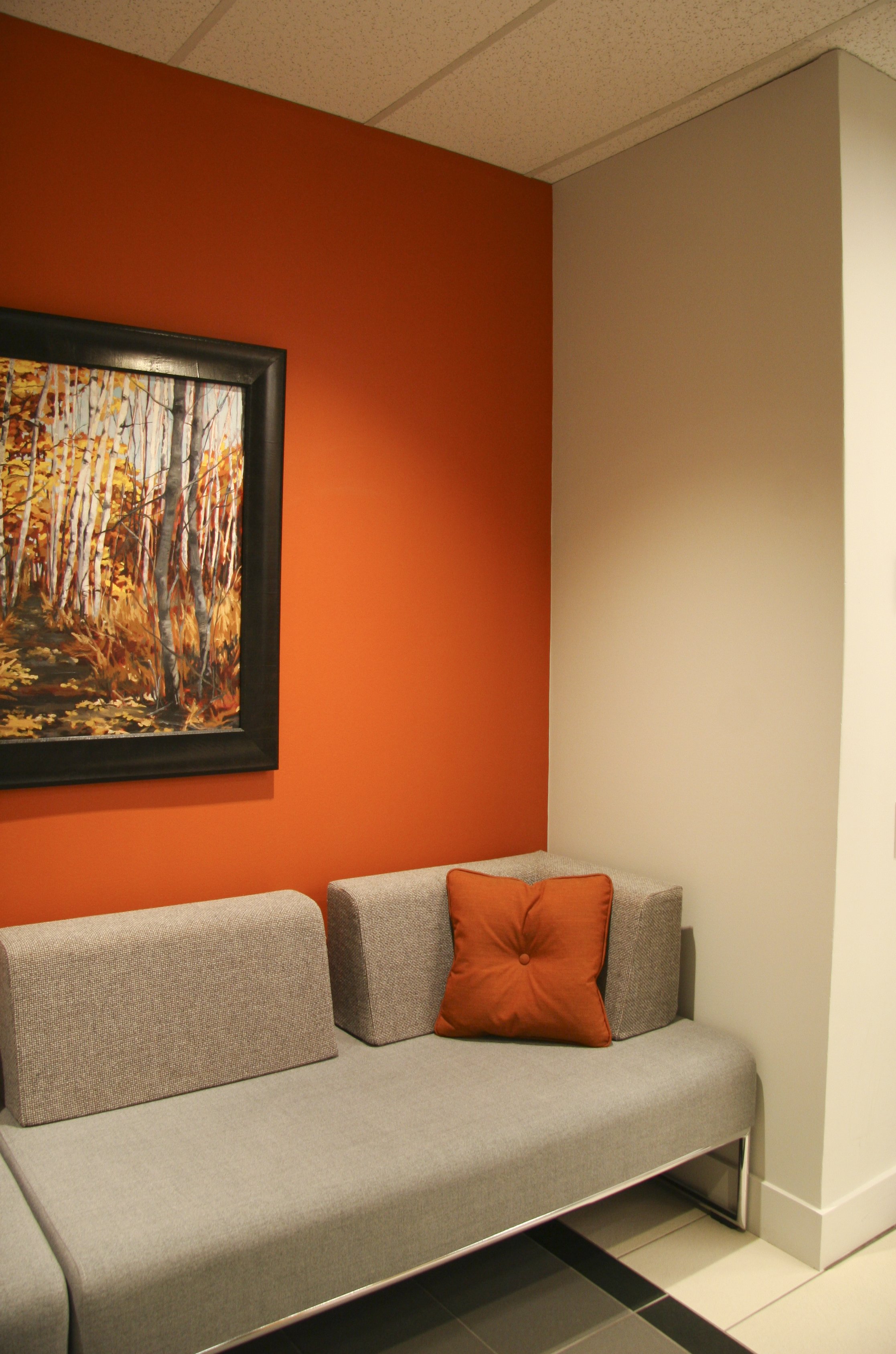 Contemporary waiting area with a gray sofa and orange accent pillow, featuring an orange accent wall displaying artwork of a forest.