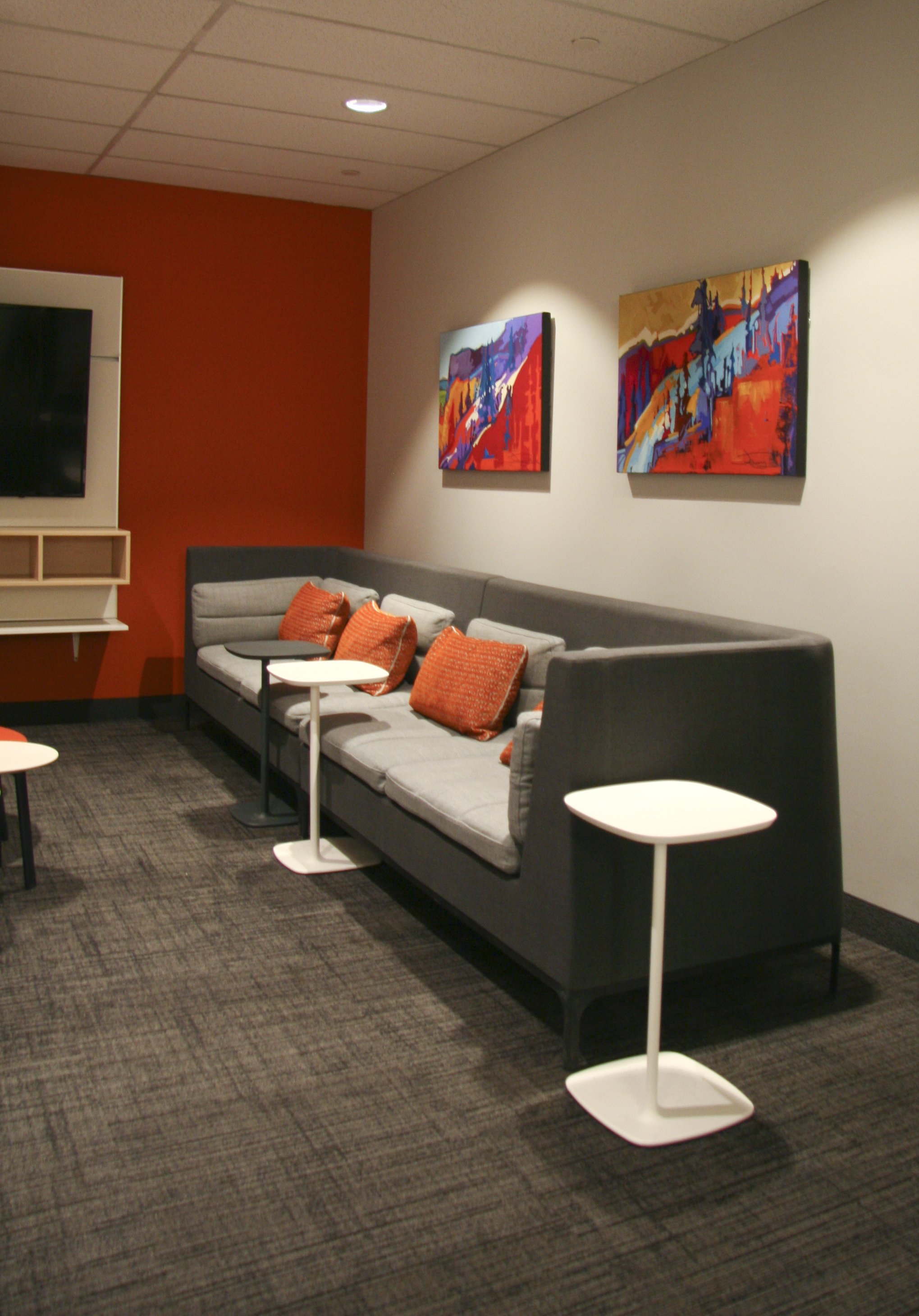 Modern office lounge with gray sectional sofa, orange cushions, small white side tables, mounted TV, abstract art, and carpet flooring.