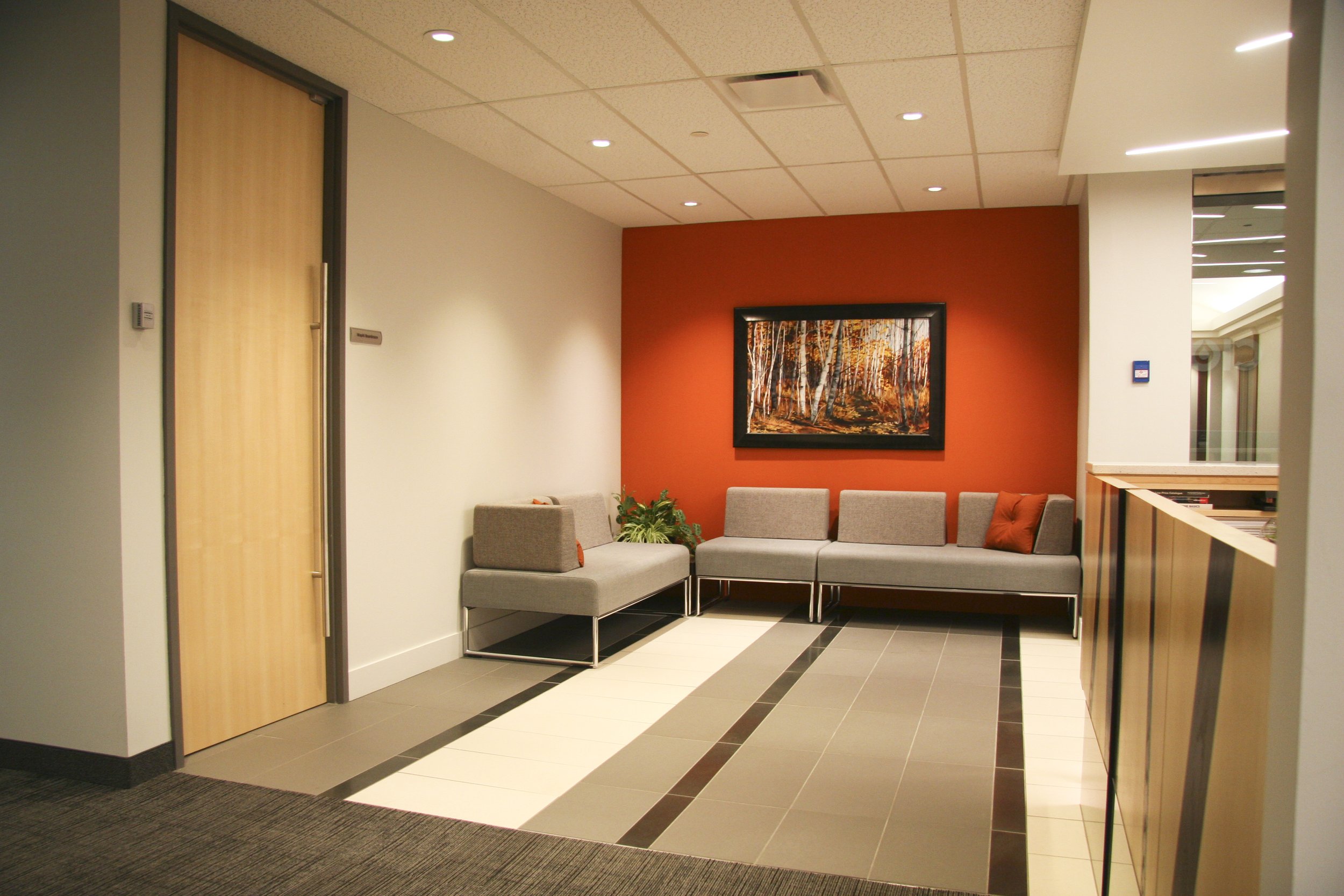 Modern waiting area with gray seating, orange accent wall, landscape painting, and neutral flooring.