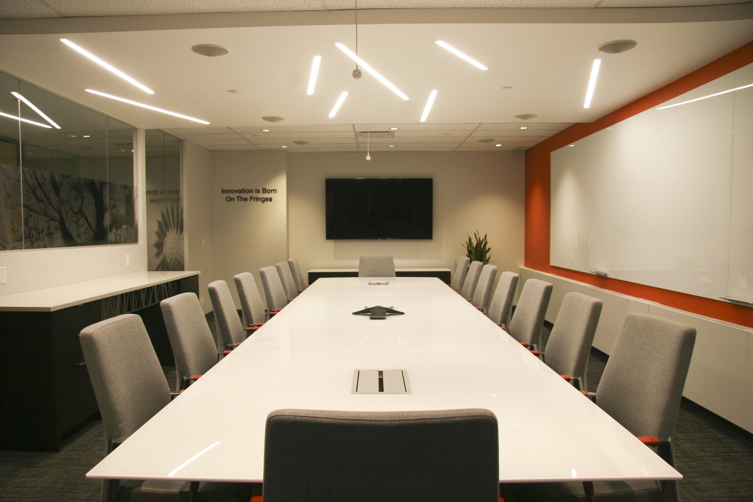 Modern conference room with a long white table, grey chairs, wall-mounted TV, and whiteboard.