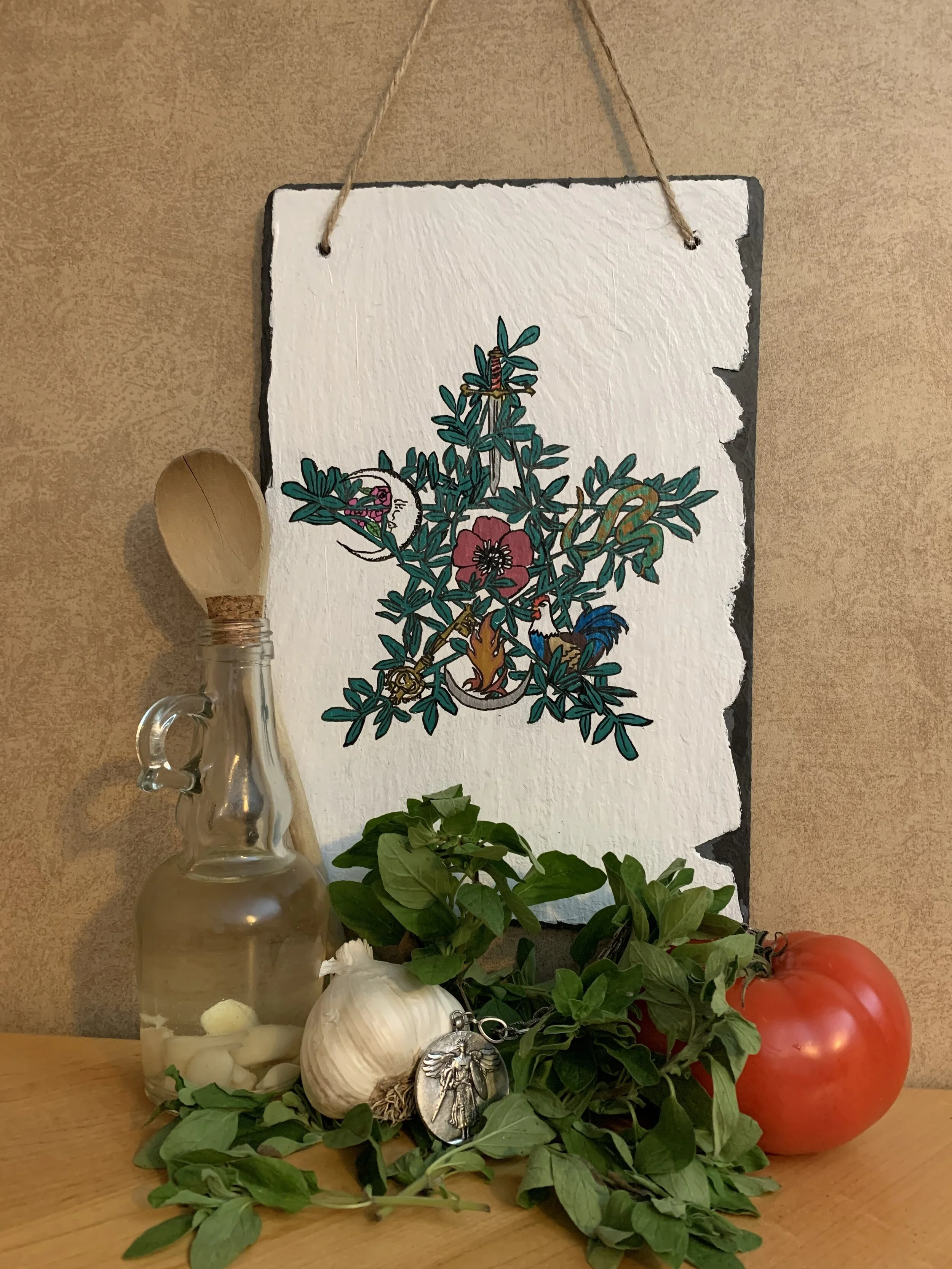 Decorative wooden sign with painted cross and birds, resting behind a glass oil bottle with a wooden spoon, fresh garlic, a sprig of green herbs, a coin pouch, and two large tomatoes on a wooden surface.