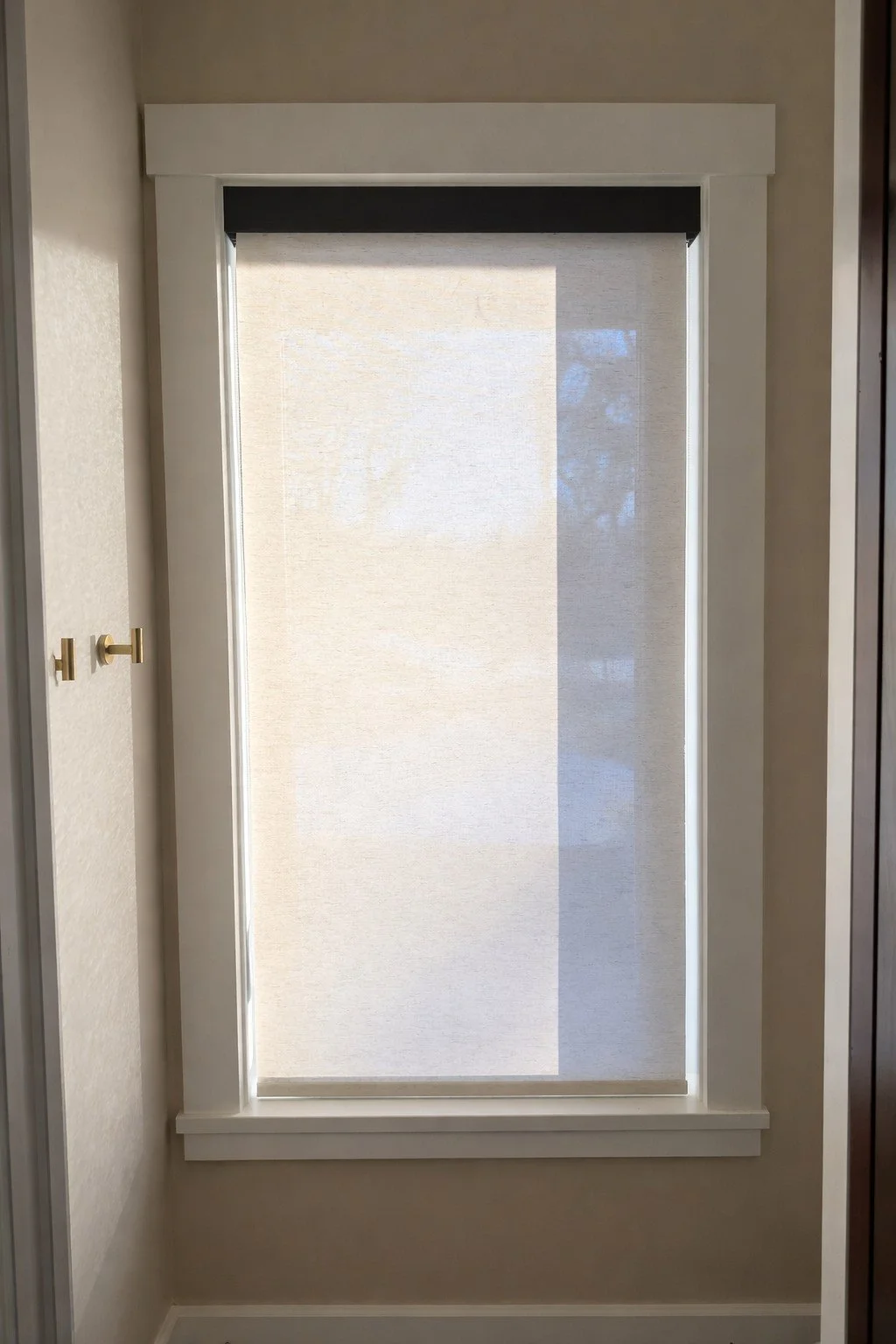 A window with a beige roller shade partially pulled down, with a dark valance at the top and white trim around the frame.