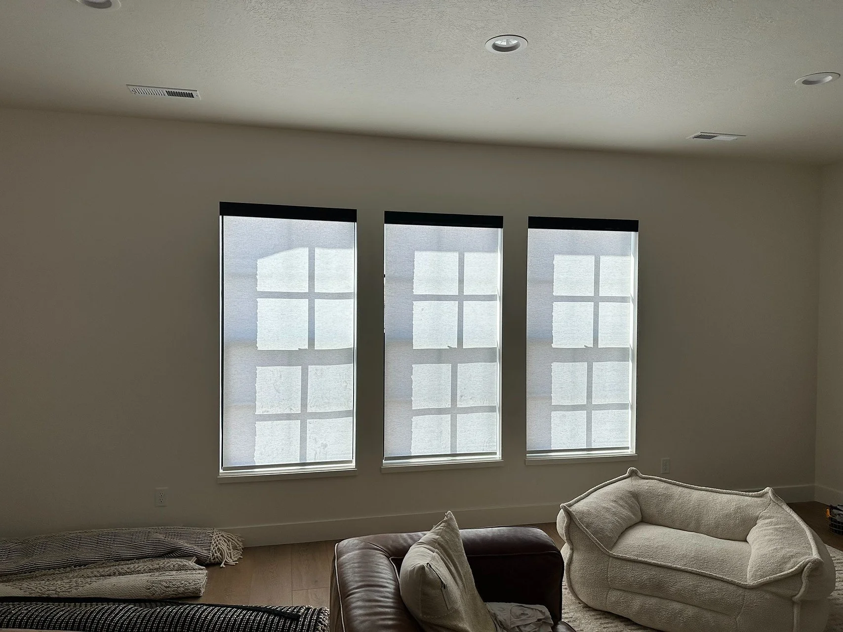 A living room with three large windows covered with white blinds, a light-colored armchair, a portion of a dark leather chair, and a pile of blankets or pillows on the floor.