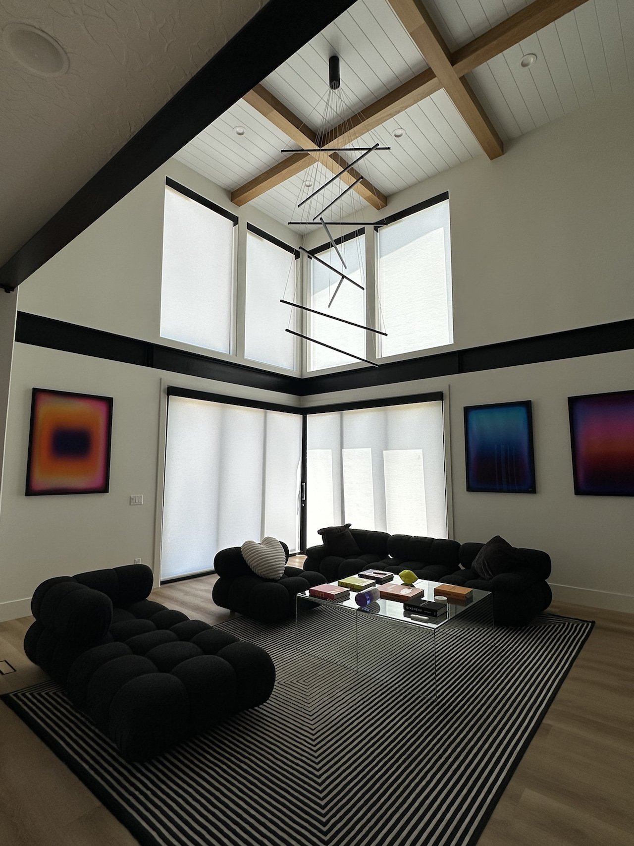 Modern living room with high vaulted ceiling, hanging abstract chandelier, large windows with white blinds, black tufted sectional sofa, striped rug, and colorful abstract artwork on white walls.