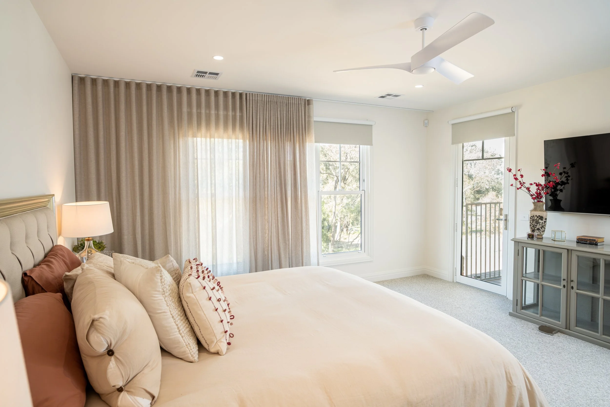 A bright bedroom with a large bed covered in beige bedding and multiple pillows, a nightstand with a lamp, and dual windows with curtains and a sliding door leading to a balcony. A TV is mounted on a stand with decorative vases and books, and a ceiling fan is overhead.