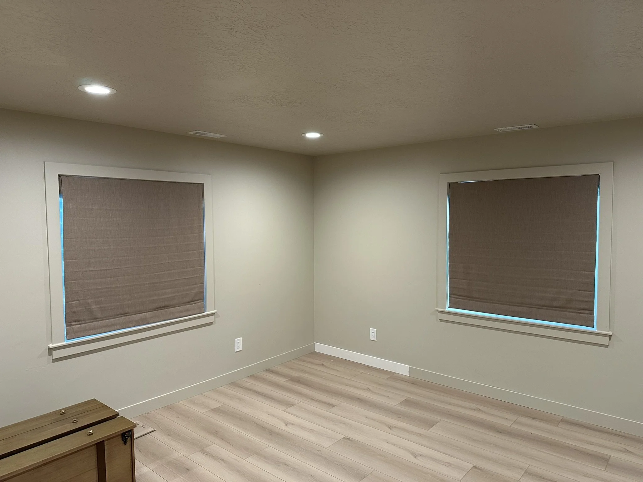 Empty room with two windows covered by brown blinds, beige walls, wooden flooring, and ceiling lights.