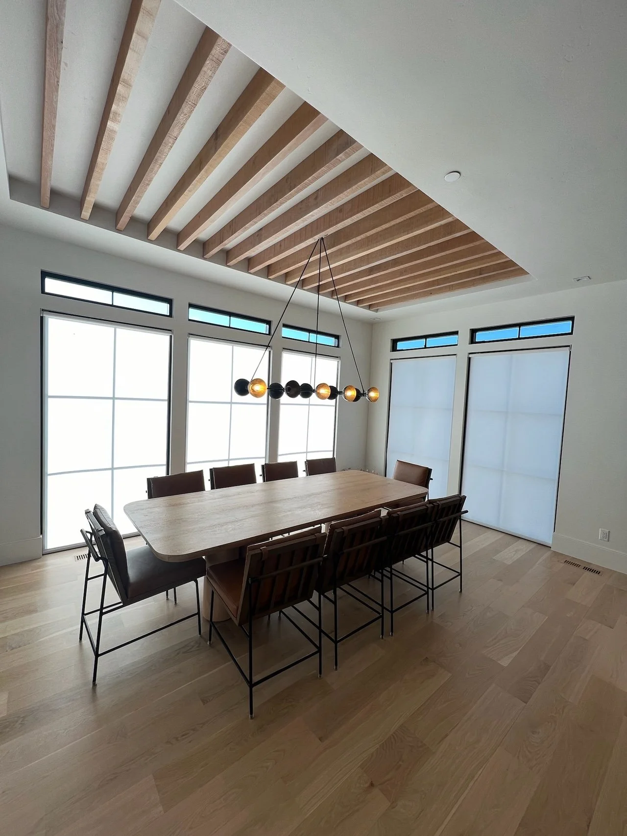 A modern dining room with large windows, a wooden table, and seven chairs. The ceiling has wooden beams and a stylish pendant light fixture with black and gold spheres. The room has light hardwood floors and white walls.