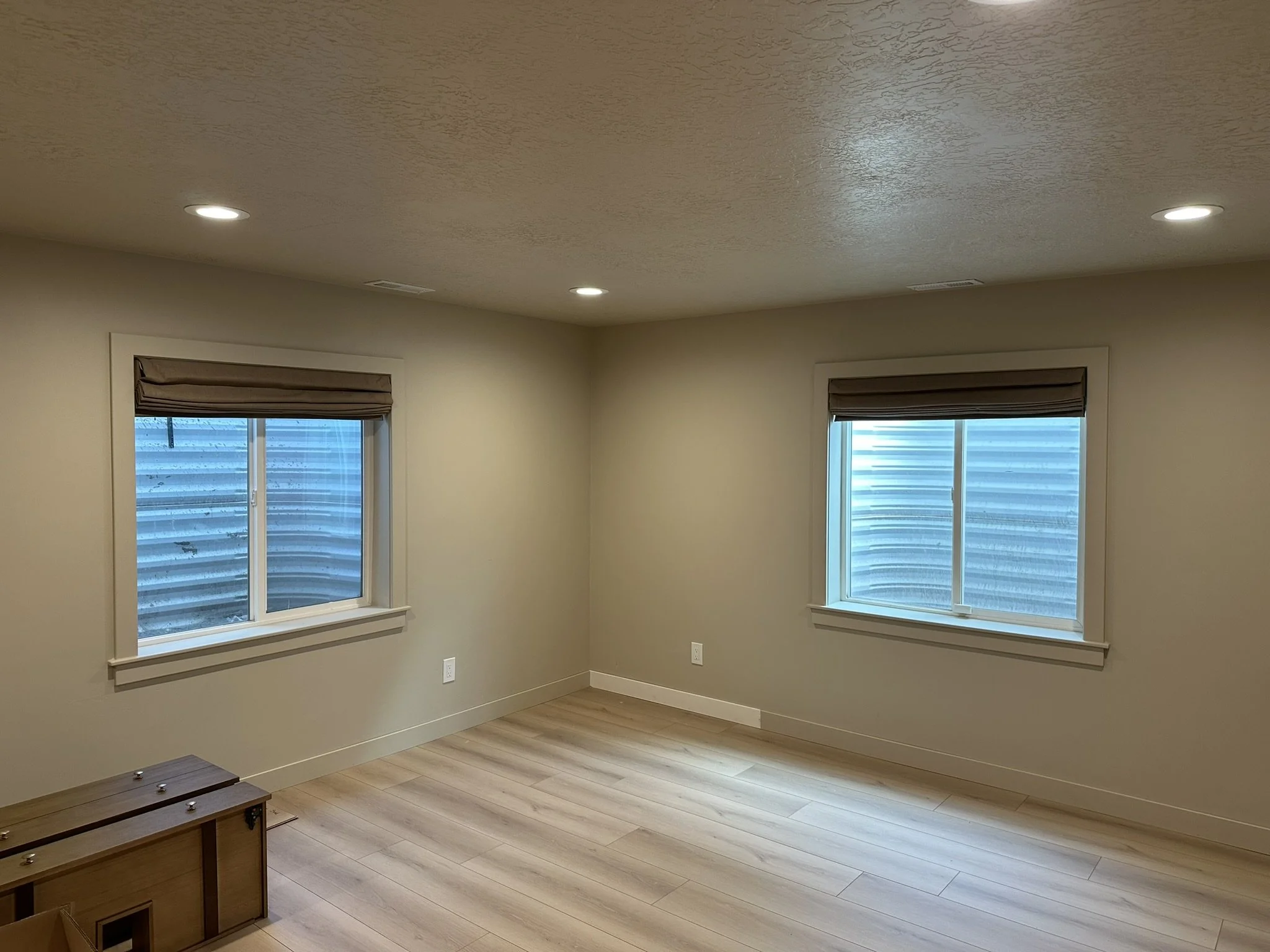 Empty room with two windows, beige walls, ceiling lights, and light-colored floor, with a small wooden piece of furniture in the corner.