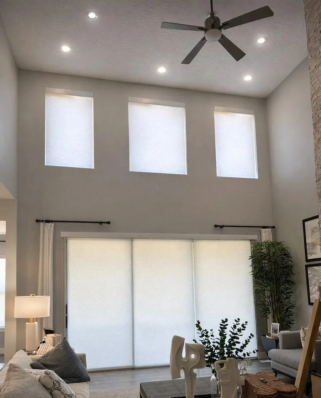 Living room with high ceiling, three small windows near the ceiling, a ceiling fan, large sliding glass door with white roller shades, a gray sofa with patterned and solid pillows, a side table with a lamp, abstract sculptures on a coffee table, hous