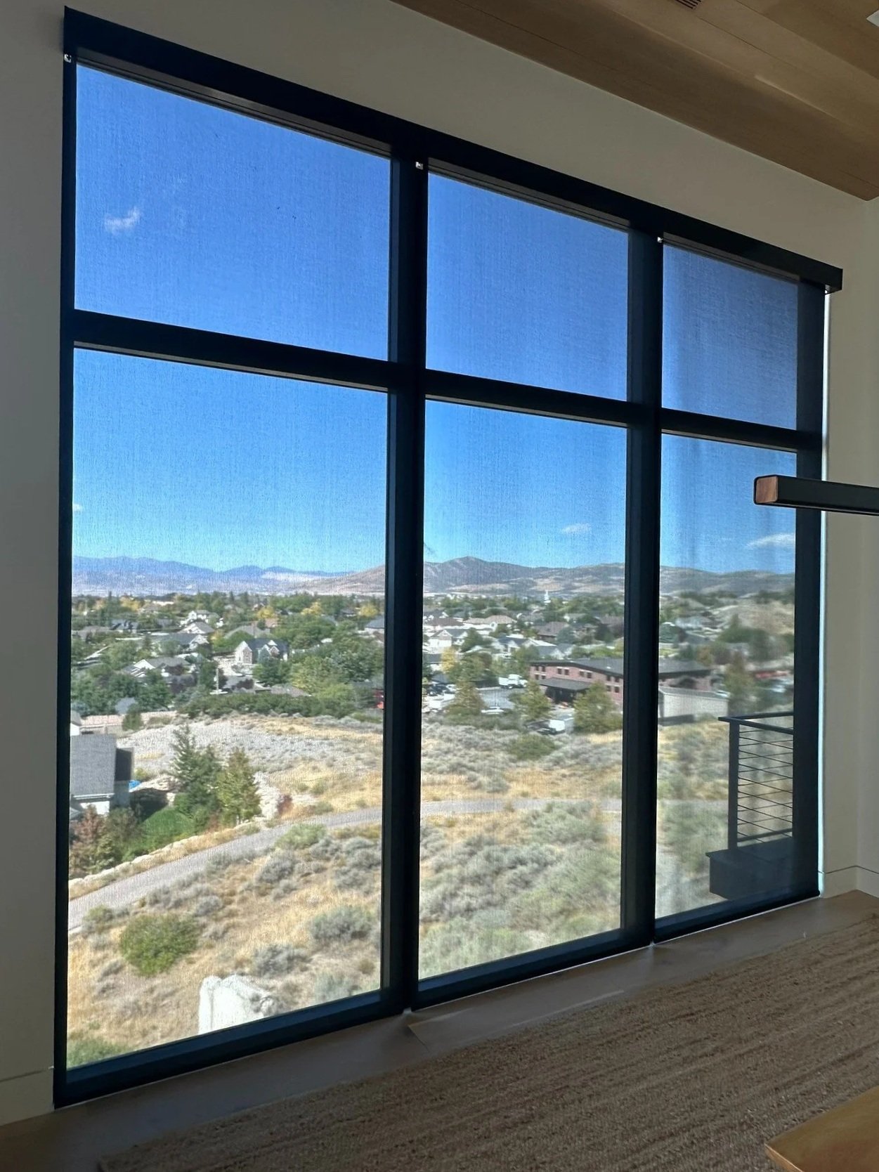Large floor-to-ceiling window with mountain and residential neighborhood view, featuring black window frames and a wooden ceiling above.