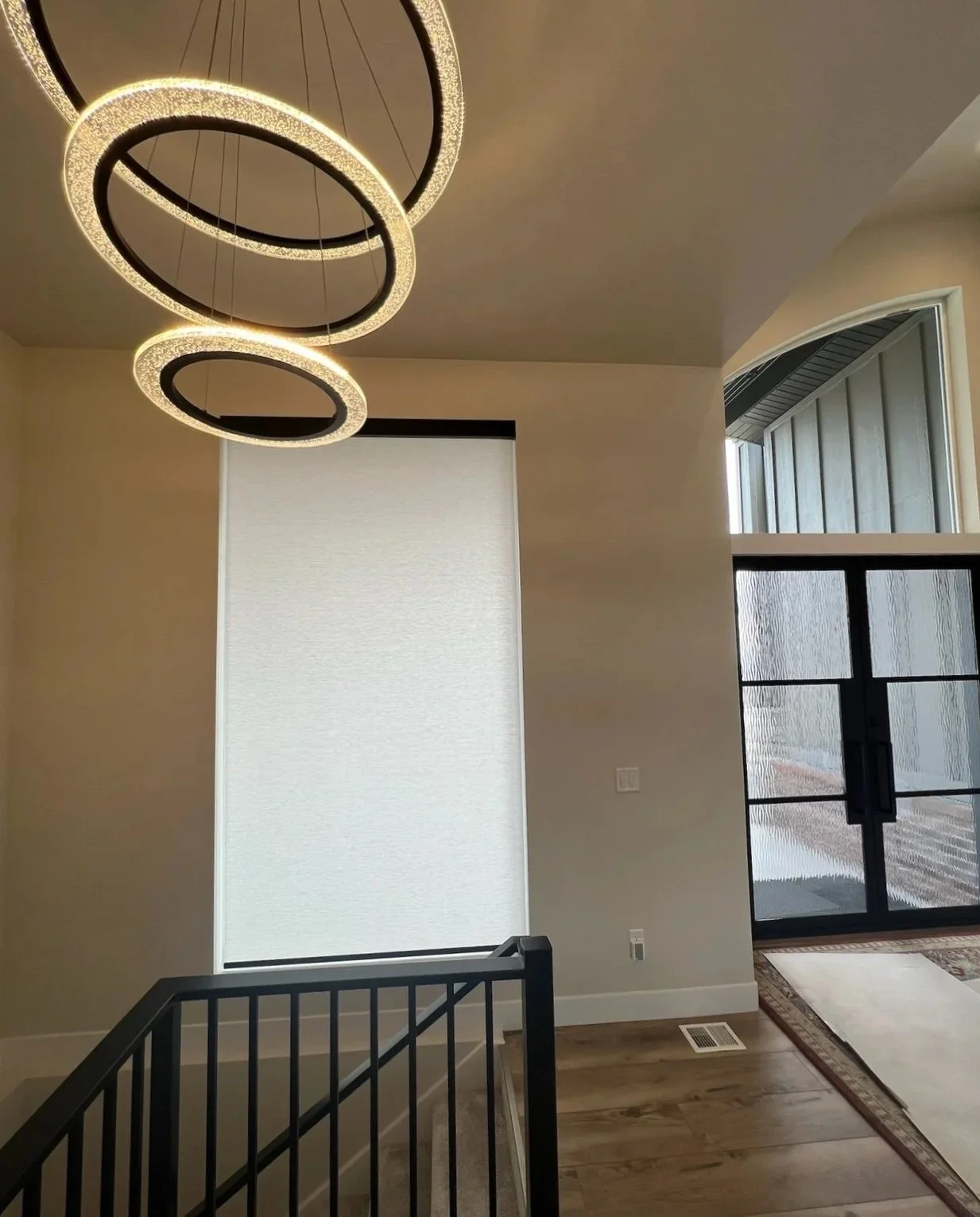 Modern interior space featuring a circular LED pendant light hanging from the ceiling, a large window with a white blind, and a glass door with a black frame next to a wooden floor and a small nest of stairs with a black handrail.
