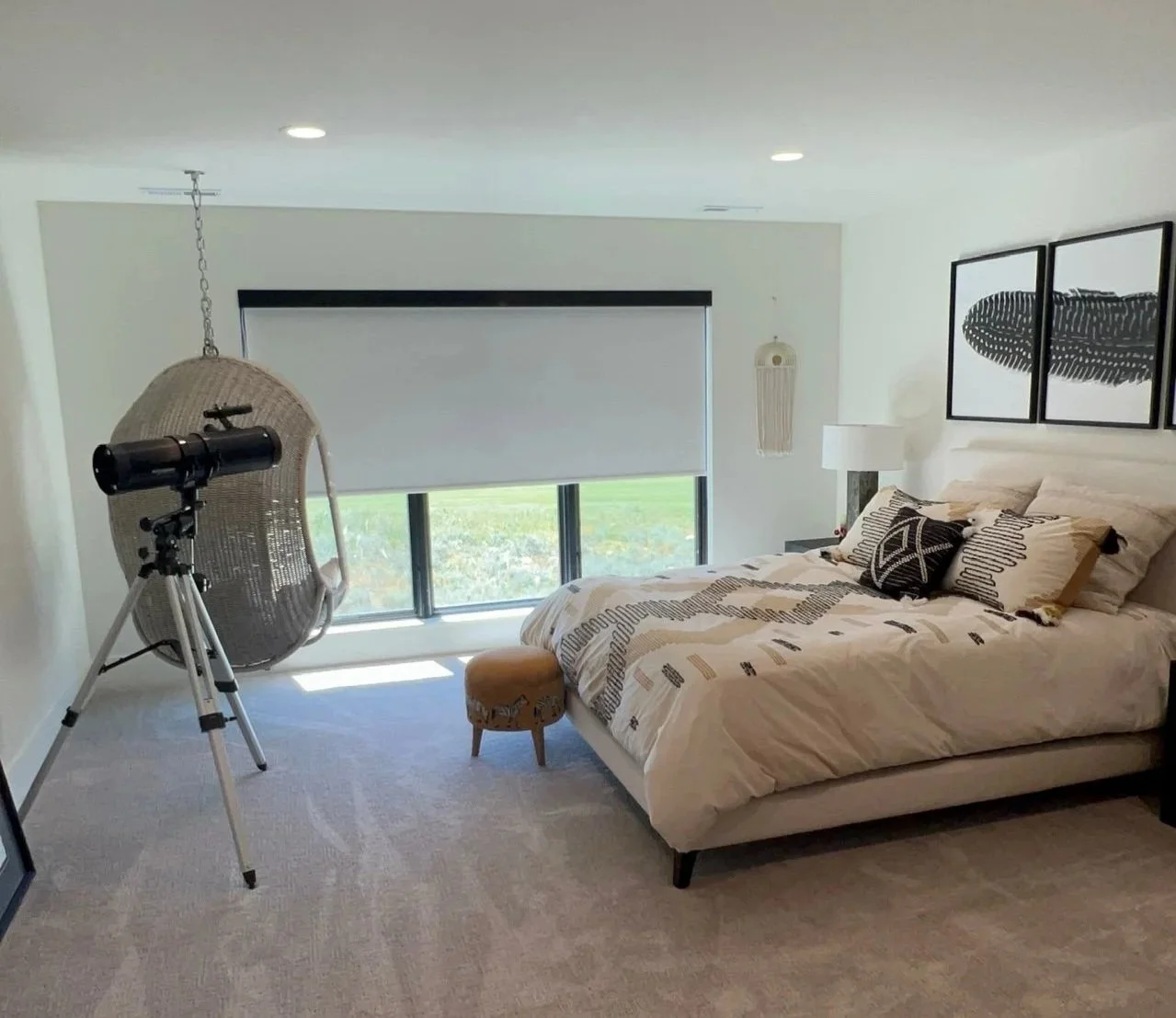 A bedroom with a large bed, black and white bedding and pillows, a small beige stool at the foot, a telescope on a tripod near the bed, a hanging chair, a window with a roller shade, and framed wall art above the bed.