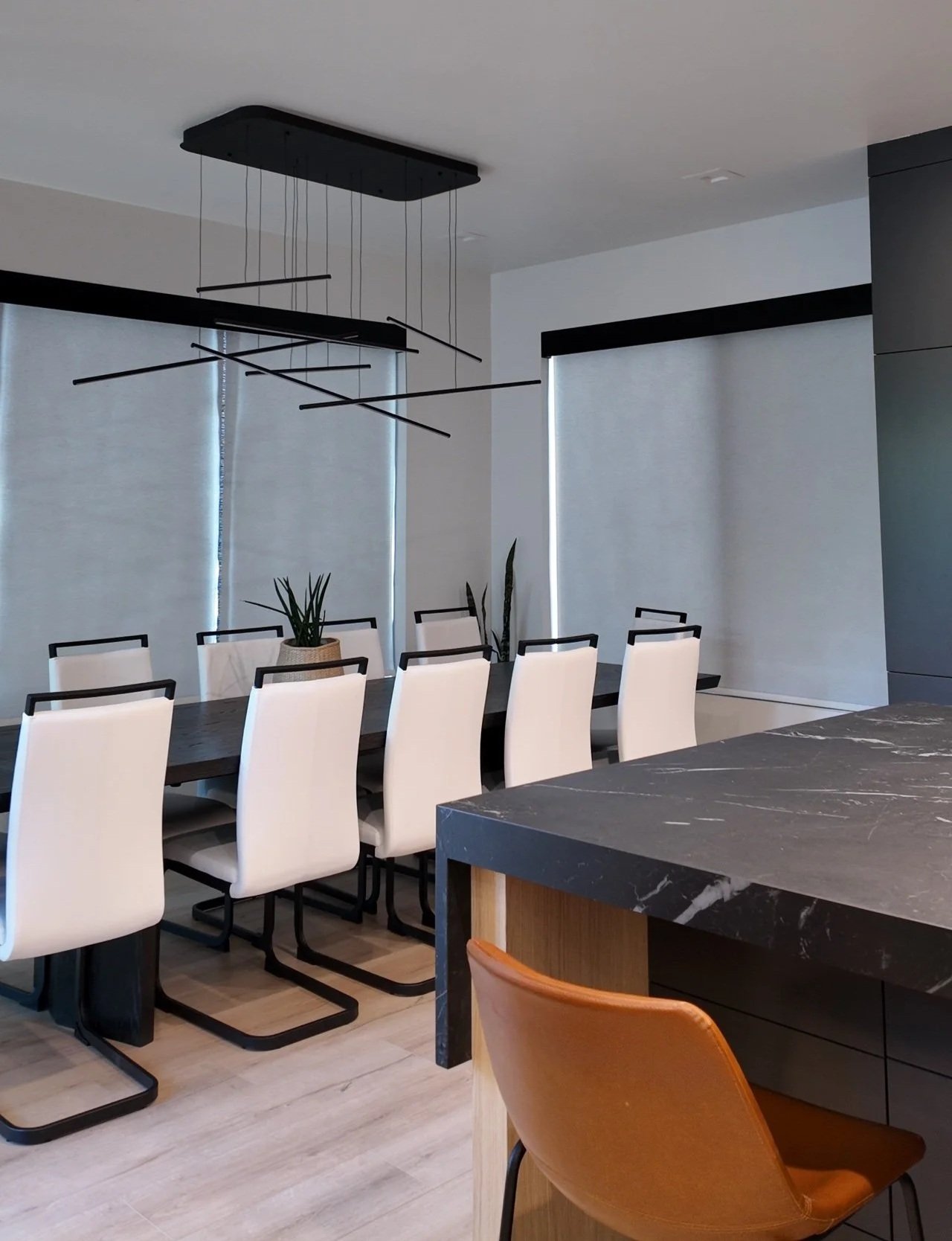 Modern dining room with a long black table, white chairs, and a black chandelier. Large windows with blinds, a marble countertop, and minimal decor.