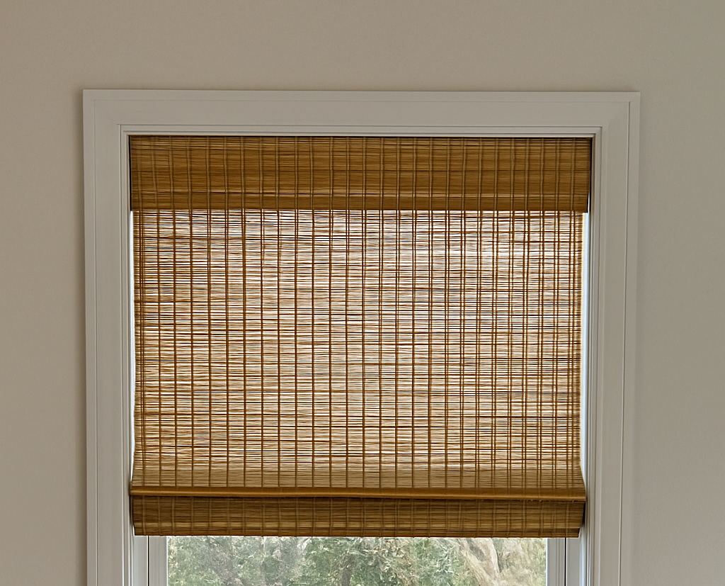 A window with a white frame and a bamboo roll-up blind partially lowered, revealing a view of trees and a wooden fence outside.