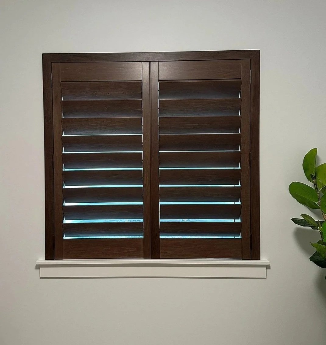 A window with wooden shutters in a room with white walls and a potted plant on the right.