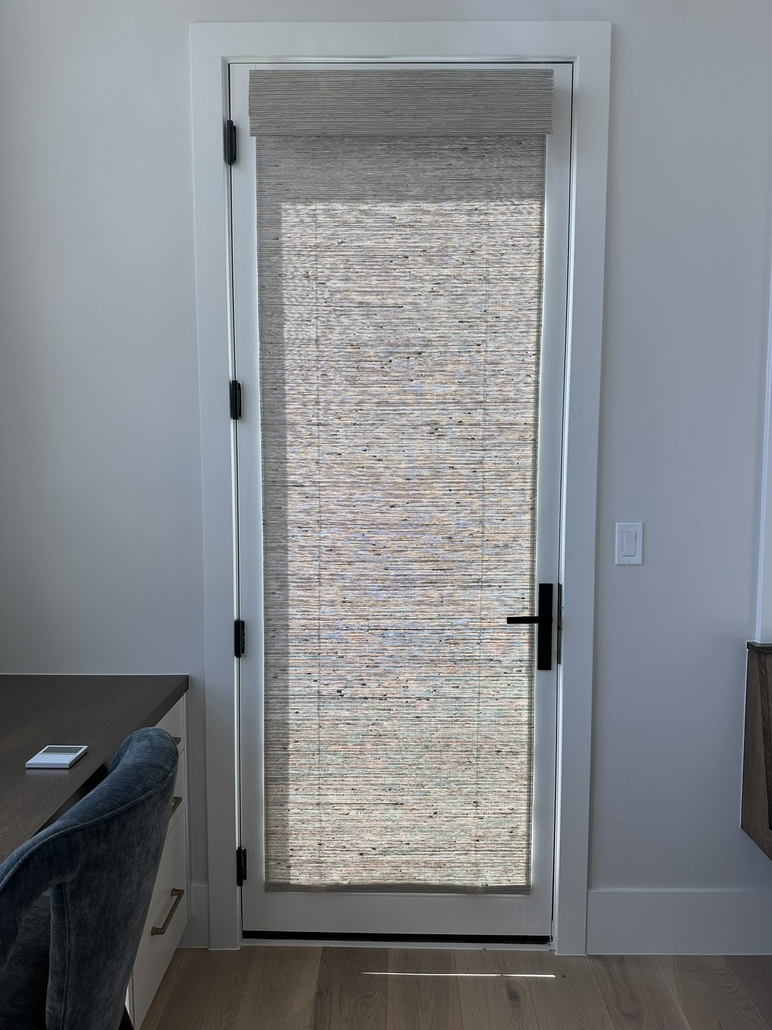 Interior view of a door with a woven blinds screen, white framing, black door handle and hinges, situated between white walls, with a light switch on the right wall, and a partial view of a desk and a blue upholstered chair on the left.