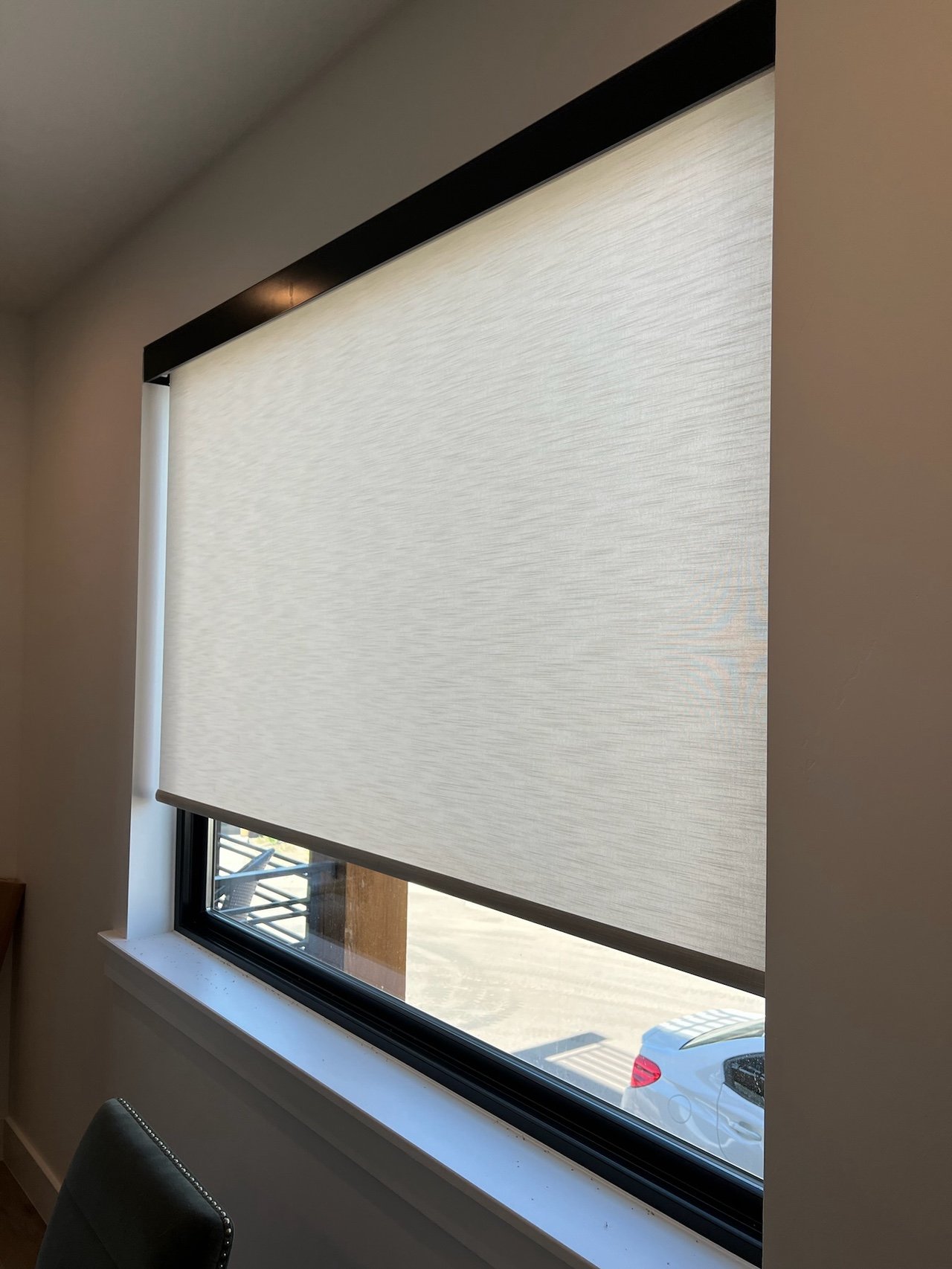 A window with a partially closed beige roller shade covering most of the glass, with a small glimpse of a parking lot and a white car outside.