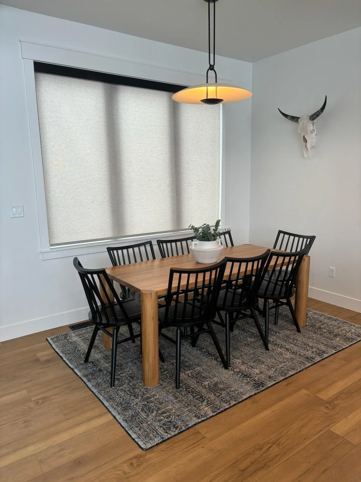 A dining room with a wooden table, six black chairs, a decorative runner, and a hanging ceiling light. There is a window with beige shades and a white skull of a bull with horns mounted on the wall.