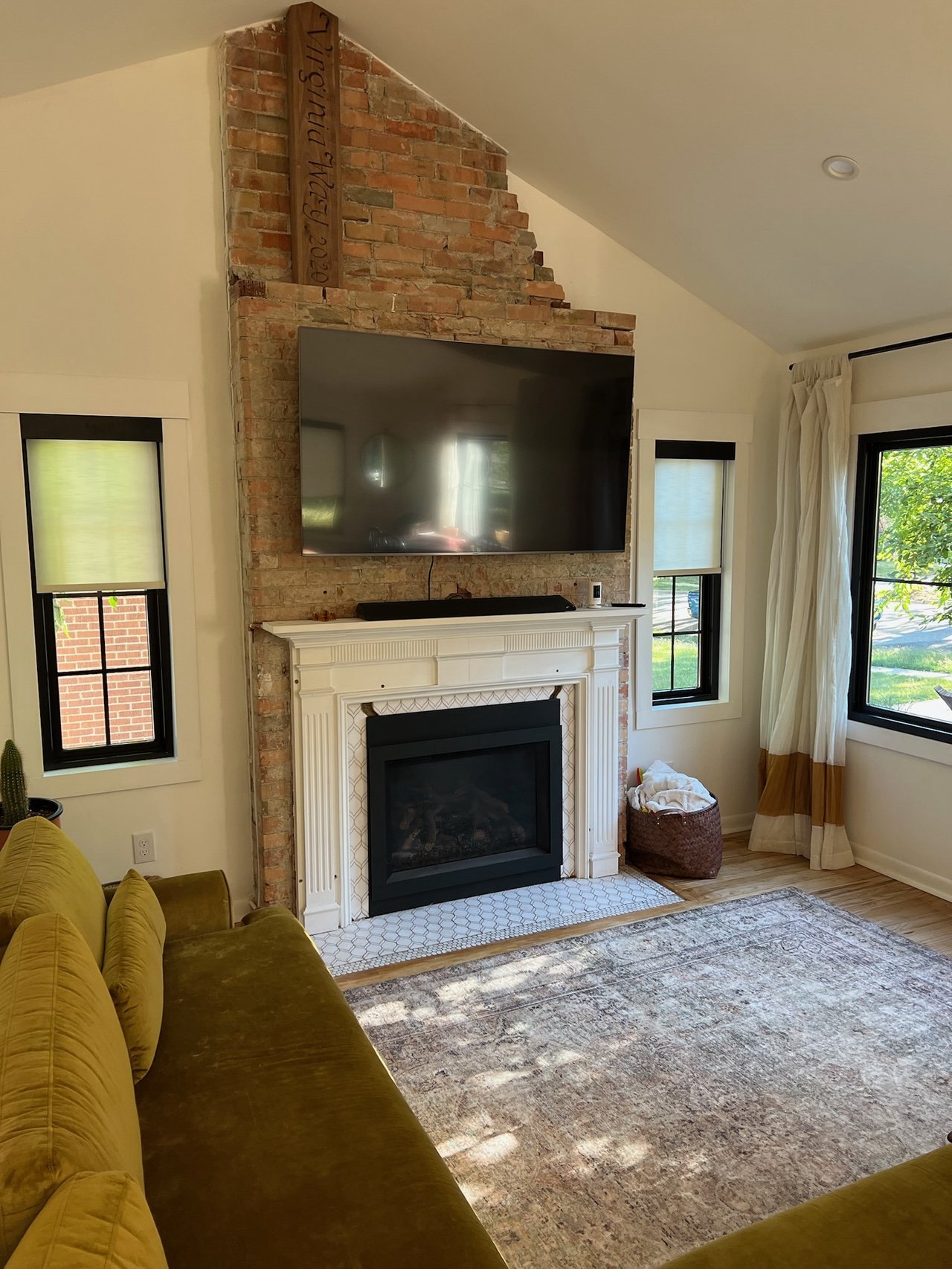 Living room with a brick fireplace, mantel, and a large flat-screen TV mounted above it. There are two windows with black frames and curtains, a yellow sofa, and a beige area rug. A basket with blankets is next to the fireplace.