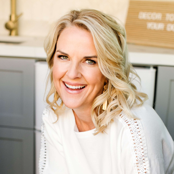 A smiling blonde woman with wavy hair, wearing a white top with decorative stitching on the sleeves, sitting in a bright kitchen with gray cabinets and a wooden sign in the background.