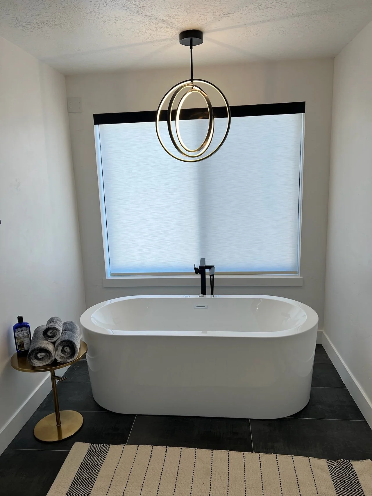 Modern bathroom with freestanding bathtub, large window with blinds, black faucet, gold and black pendant light, towel rack with rolled towels, and a beige rug.