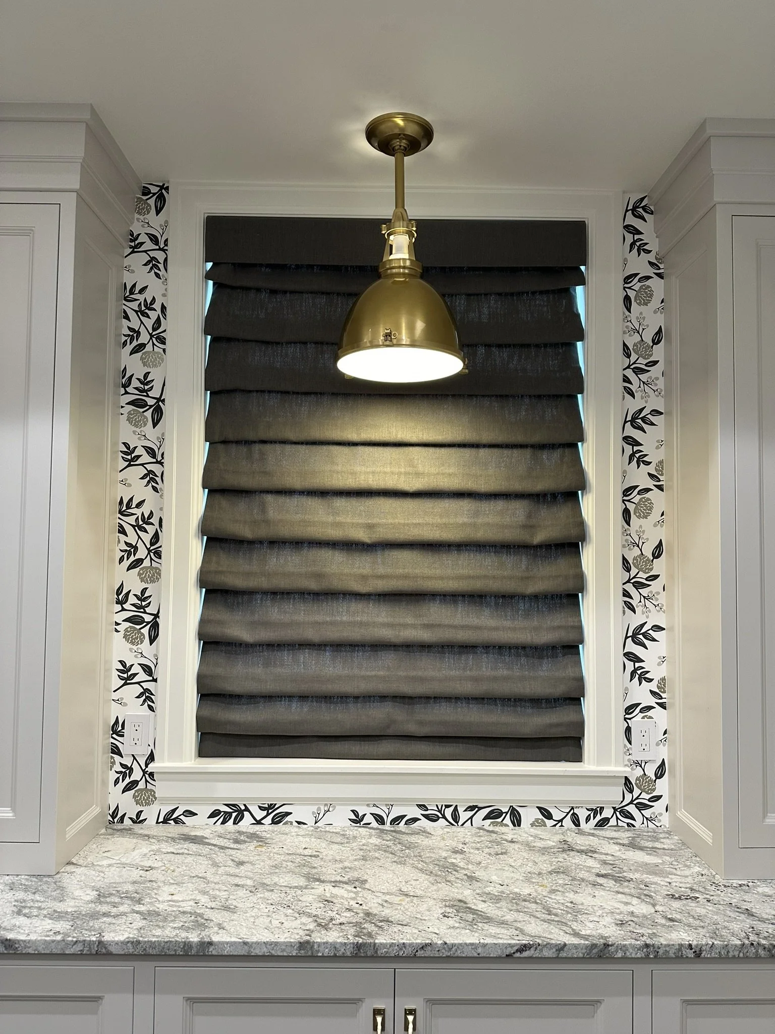 Window with black Roman shades, white trim, floral wallpaper border, above a gray marble countertop, with gold pendant light hanging below the ceiling.