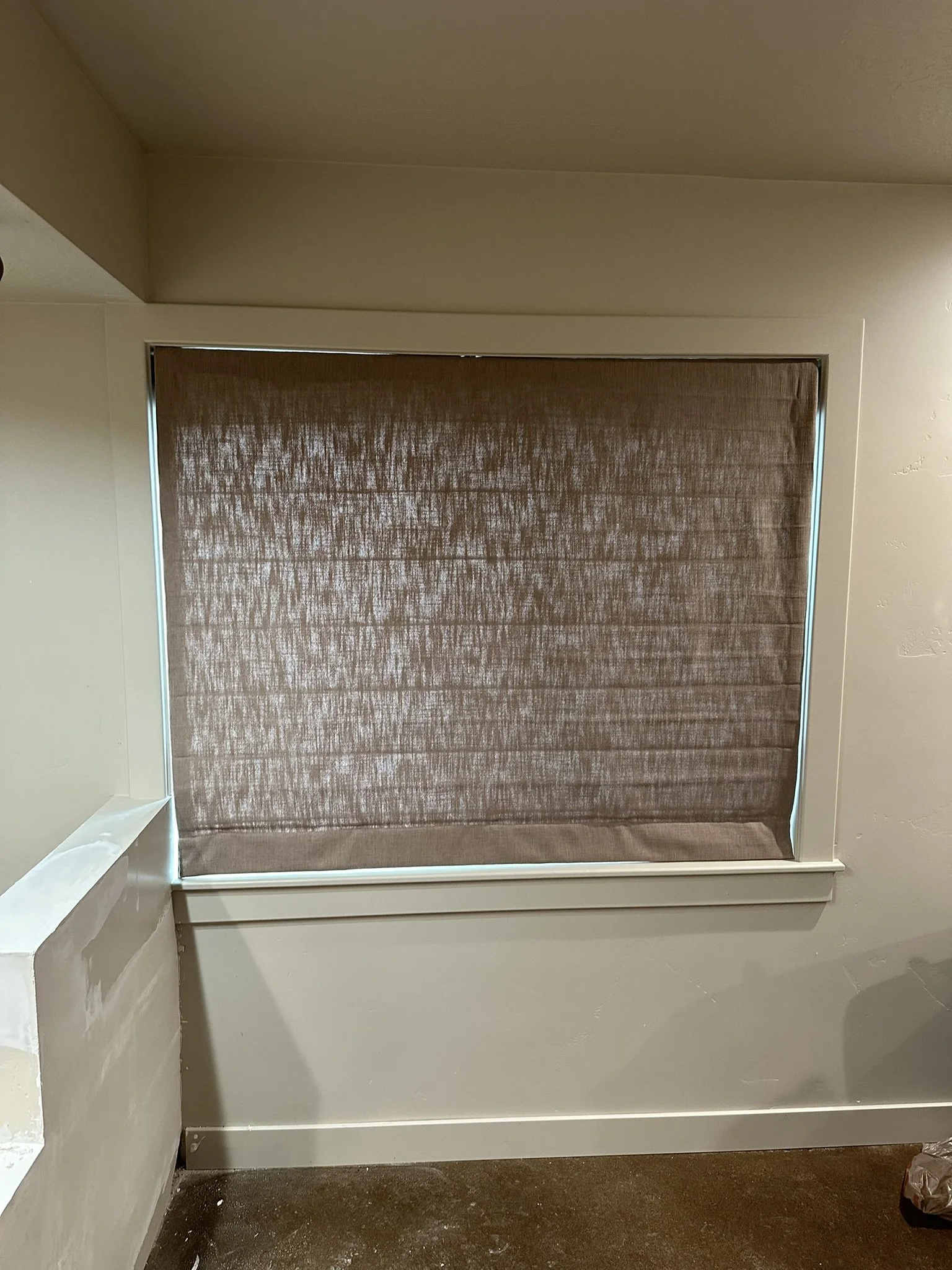 A window covered with a brown textured roller shade in a room with beige walls and dark brown carpet.