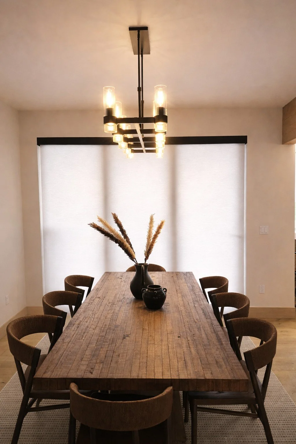 A modern dining room with a long wooden table, eight brown chairs, a black vase with pampas grass, a small black jar, a large window with white blinds, and a contemporary black chandelier with exposed bulbs.