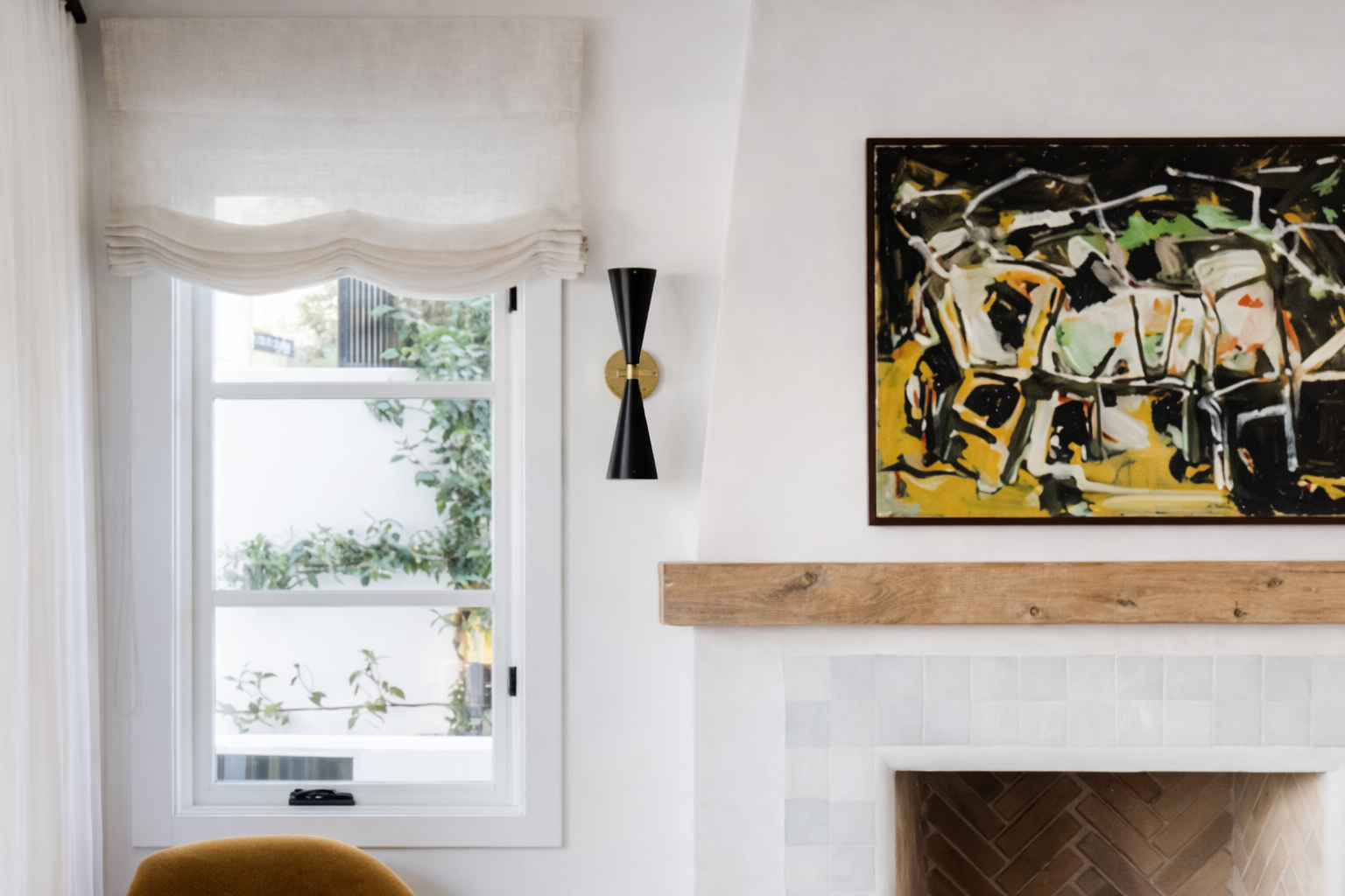 A bright living room with a window covered by a white roman shade, a black and gold wall sconce, an abstract painting with black, yellow, green, and white colors, a wooden mantle, a fireplace with brick interior, and a yellow upholstered chair partia