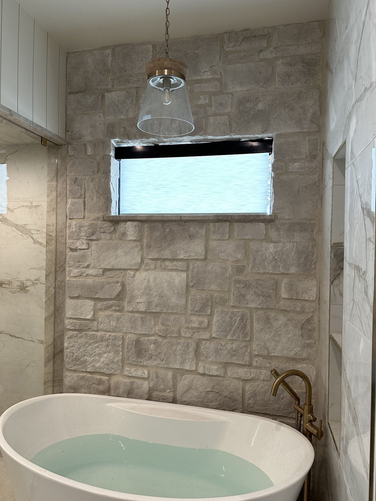 A bathroom with a freestanding bathtub filled with water, a stone wall, a small horizontal window, and a pendant light fixture hanging from the ceiling.