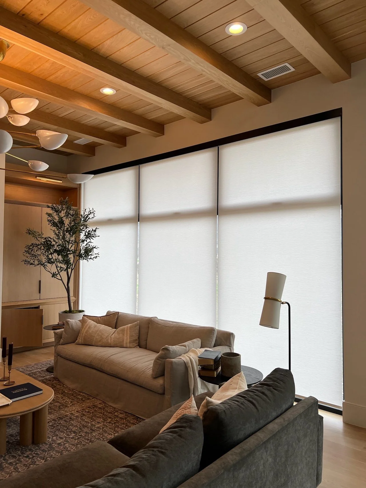 A cozy living room with a beige sofa, a dark gray armchair, a coffee table, and large windows with white blinds. The room features a wooden ceiling with exposed beams and a modern chandelier.