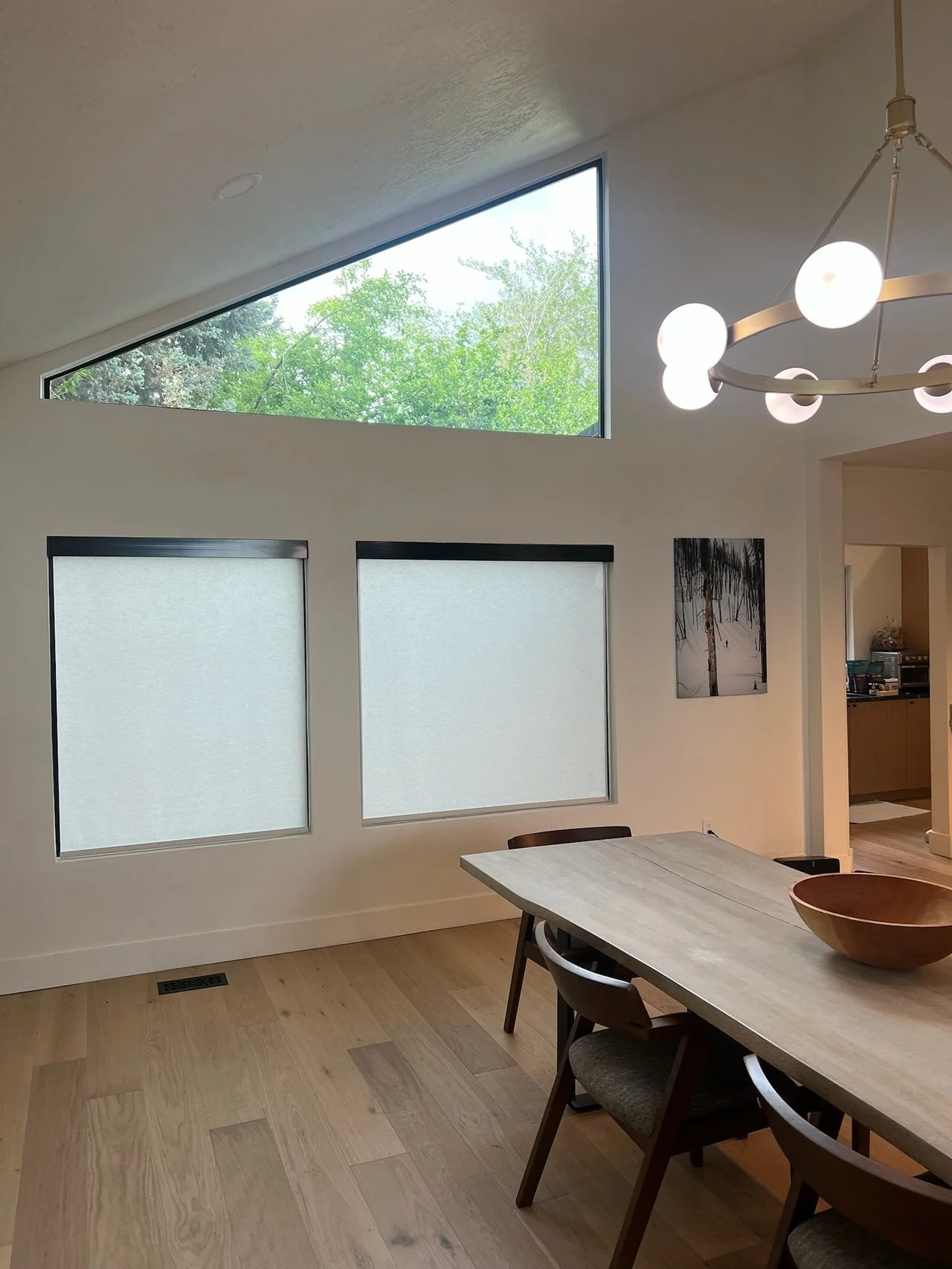 Interior view of a dining room with a wooden table and chairs, modern hanging light fixture, two frosted windows, and a large triangular window. Tree canopy visible outside the window.