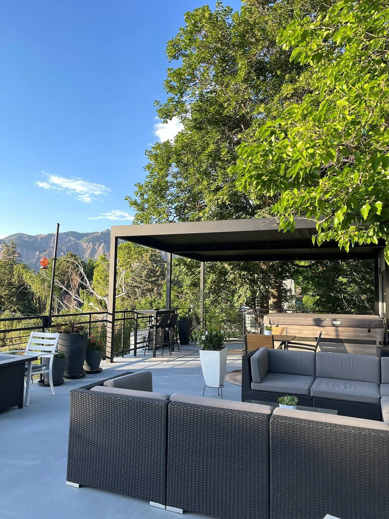 A rooftop outdoor patio with outdoor furniture including sofas, a table, bar stools, potted plants, and a shaded pergola, surrounded by green trees and mountain views in the background under a clear blue sky.