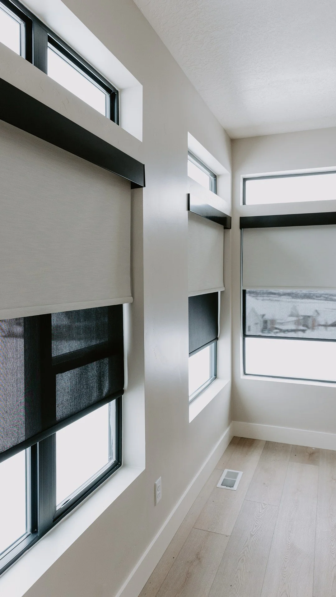 Room with large windows covered by roller shades, white walls, light wood flooring, and a floor vent.