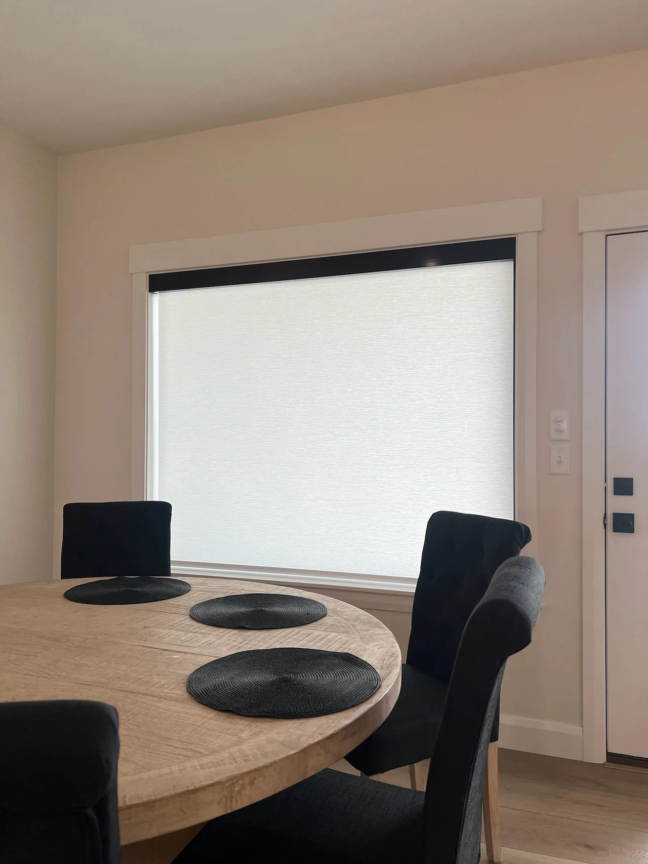 A dining room with a round wooden table, four black chairs, and a window with closed blinds.