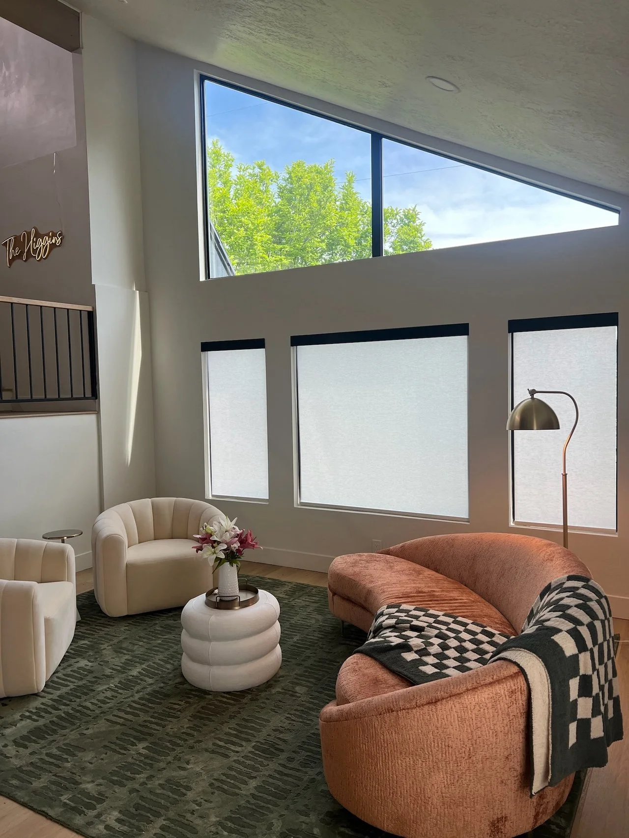 Modern living room with large triangular upper window, including three rectangular lower windows with frosted glass, featuring a peach curved sofa, two round beige armchairs, a white textured side table with a floral vase, a green patterned rug, and 