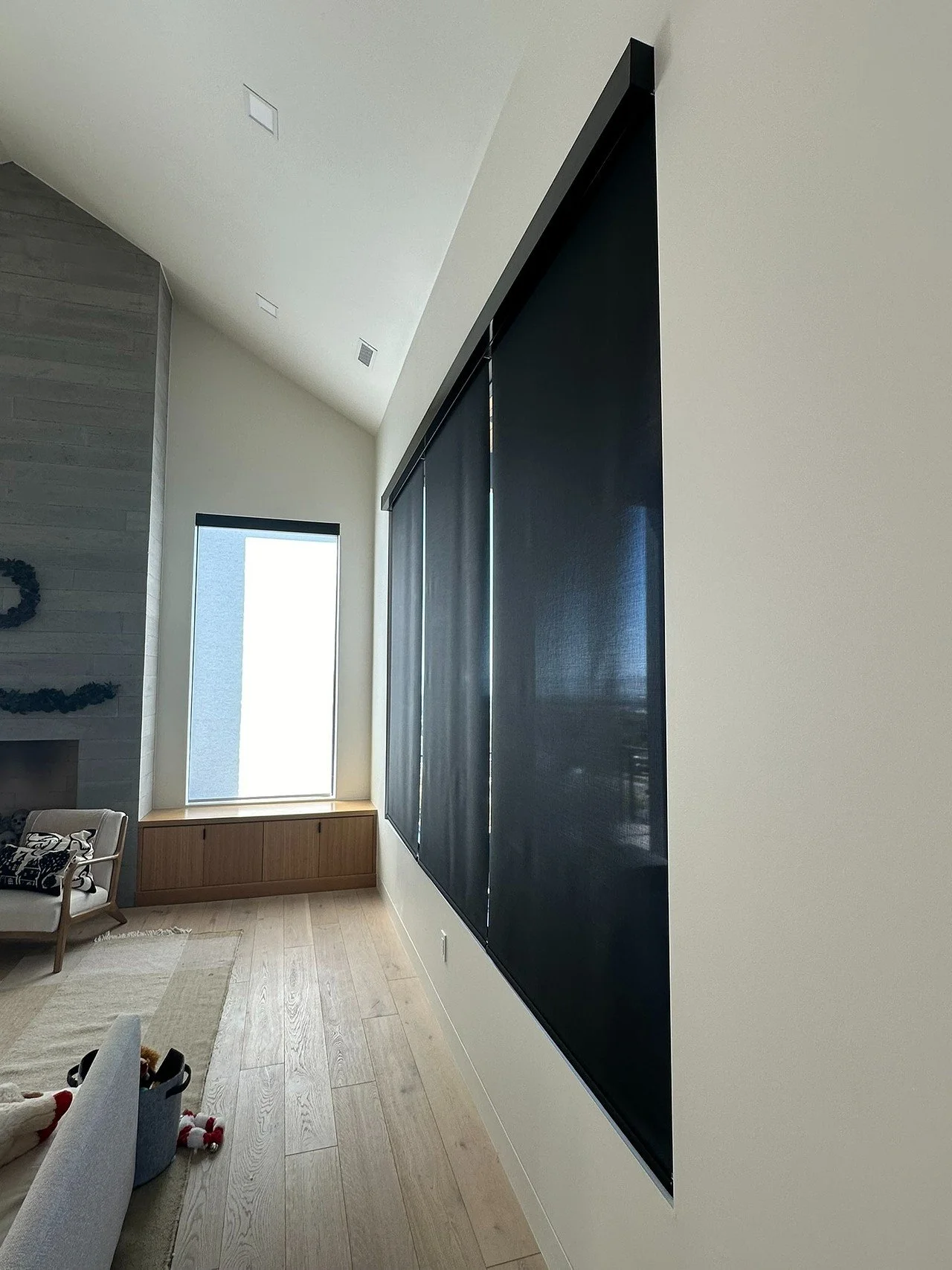 Modern living room with a sloped ceiling, large black window shades, a tall window with blinds, a fireplace, and a beige armchair.