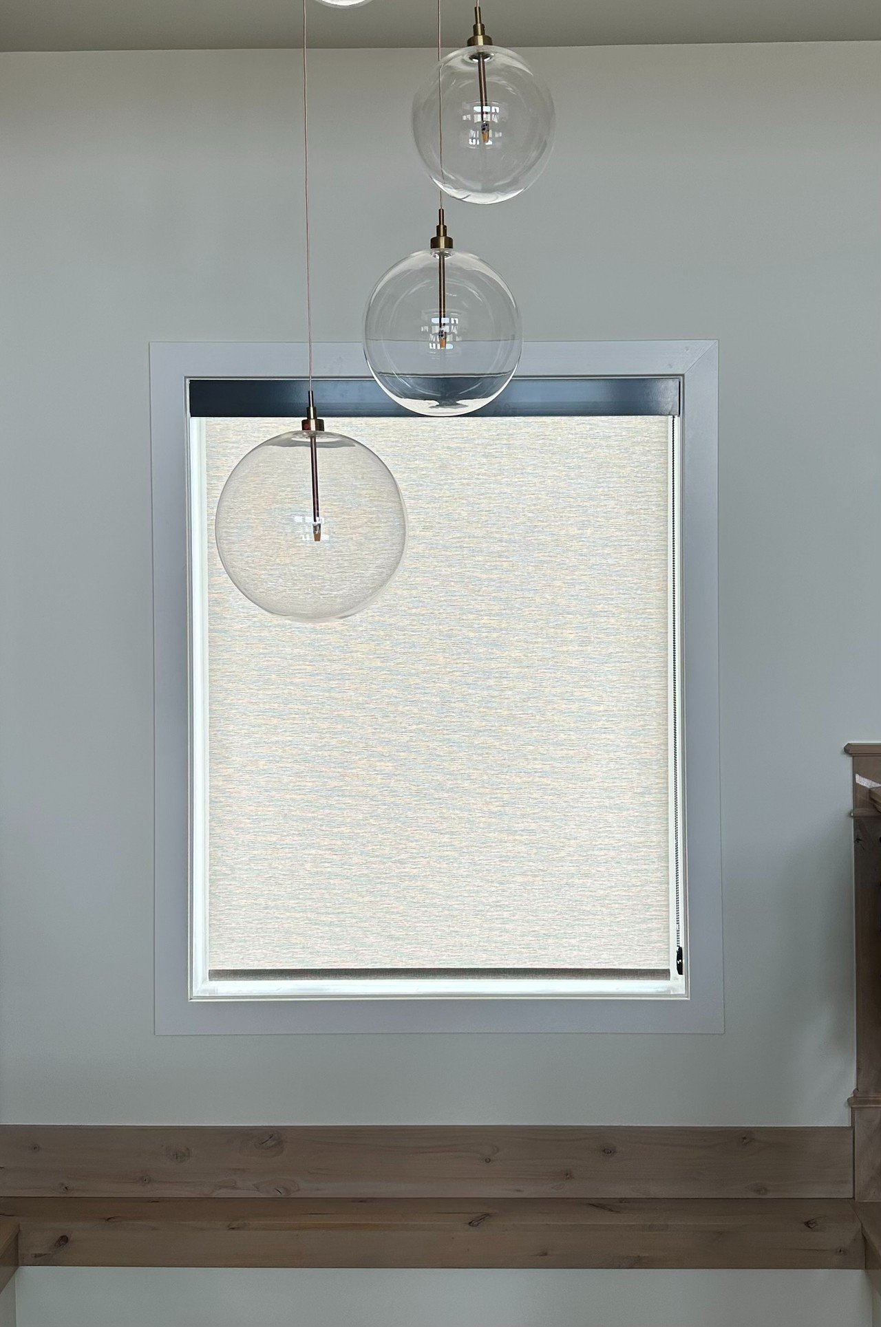 Three hanging glass pendant lights in front of a large window with a beige roller shade, in a room with wooden flooring and a light green wall.