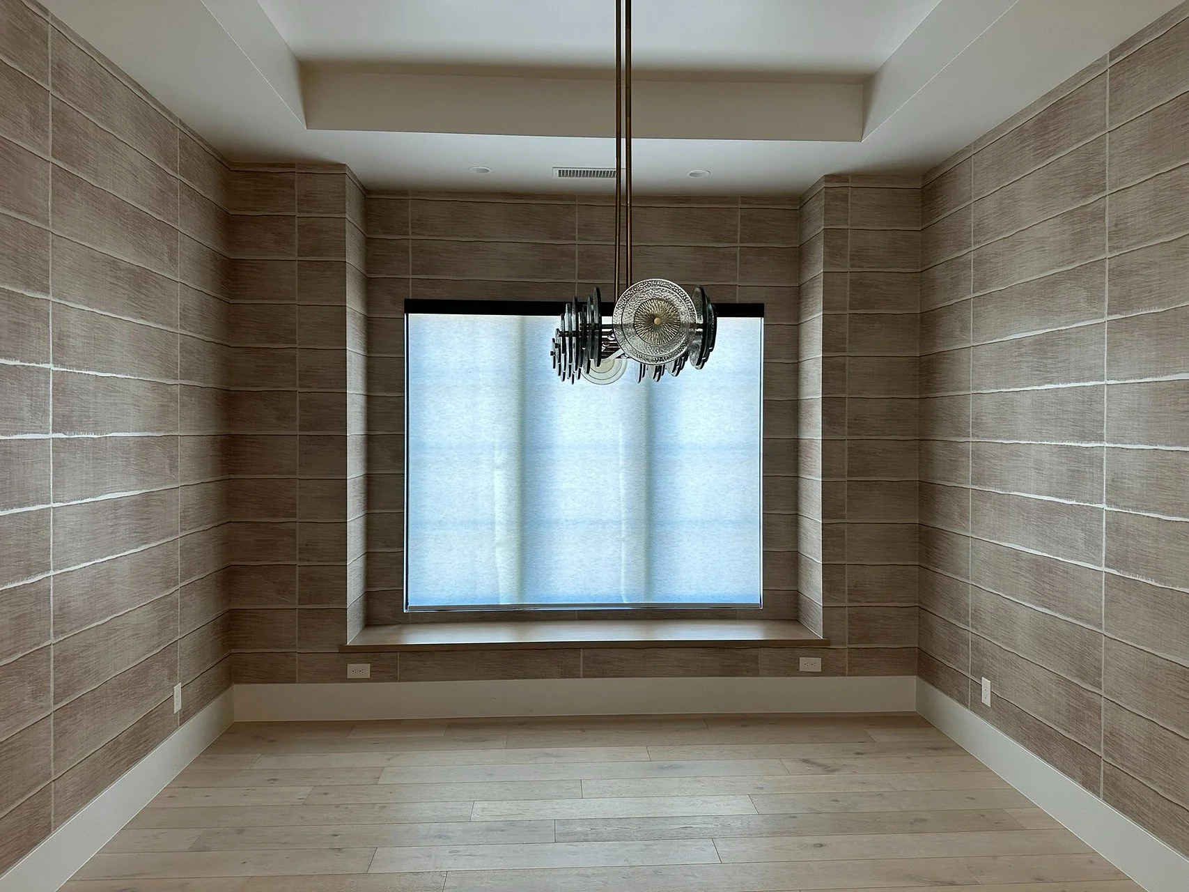 Empty room with light wood flooring, beige brick-textured walls, large window with frosted glass, and a modern black and silver chandelier hanging from the ceiling.