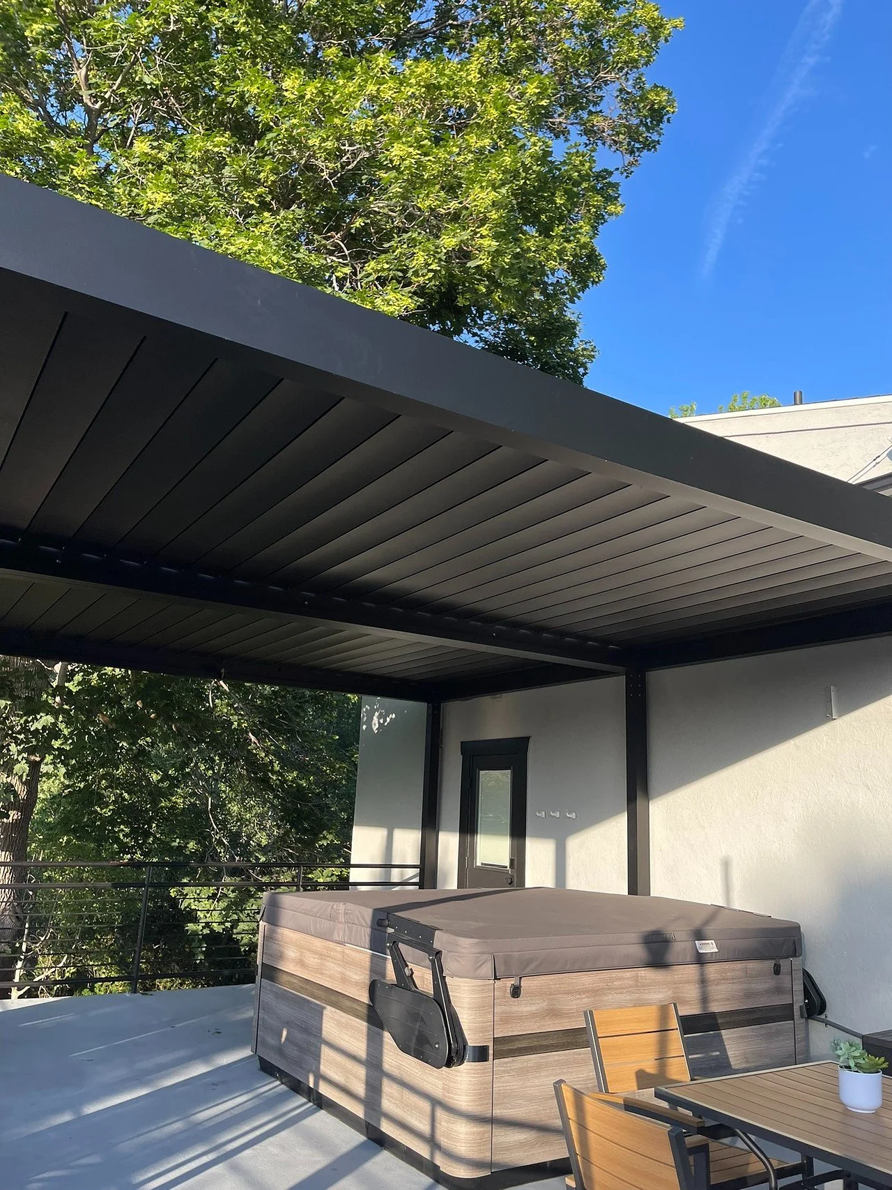 Outdoor patio area with hot tub, wooden table, and chairs beneath a black metal pergola, with a view of green trees and a blue sky.